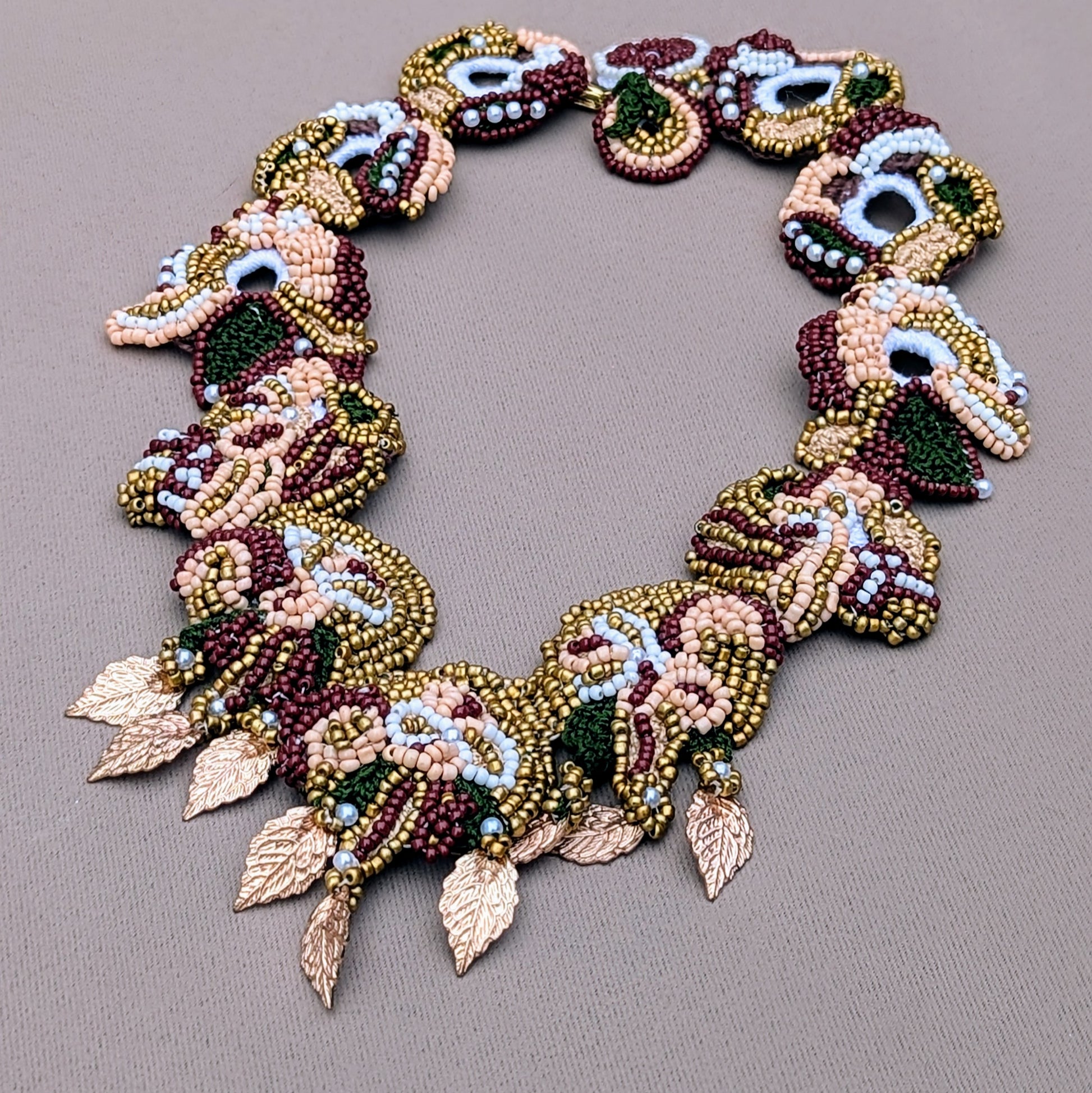 An artistically handcrafted, one of a kind necklace available exclusively from the sustainable UK luxury brand Mayaani Jewellery. This artisan piece features intricate beadwork in traditional shades of gold, red, green and ivory, arranged in an elaborate pattern of swirling designs. Adorned with delicate gold leaf accents, the necklace showcases exceptional craftsmanship and is designed for those seeking a unique and luxurious accessory. Perfect for special occasions or bridal wear.