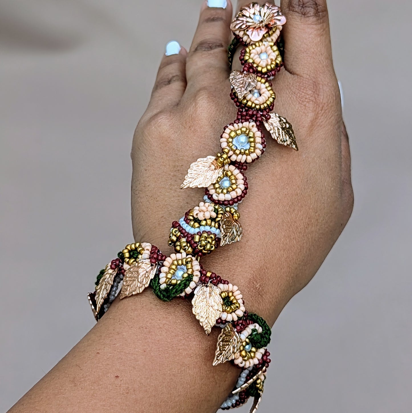 This Indian hand bracelet (haath phool) is a beautifully crafted piece that blends traditional bridal charm with modern design, exclusive to sustainable brand Mayaani Jewellery UK. Featuring red, green, gold, and ivory pearl seed beads, it can be styled as a bracelet or adjustable ring, adorned with rose gold leaf and flower accents. The only piece you need for weddings, occasions and everyday.