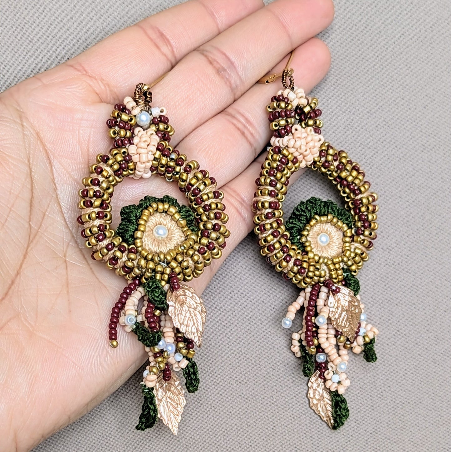 A unique pair of Indian bridal earrings exclusively handcrafted by the sustainable UK brand Mayaani Jewellery, displayed on a white peacock shaped trinket dish. The earrings feature intricate red, green and gold beadwork with circular patterns, embellished with leaves and delicate crochet detailing. These one of a kind earrings reflect traditional Indian bridal design, combining exceptional craftsmanship with high quality materials making them an elegant and timeless accessory.