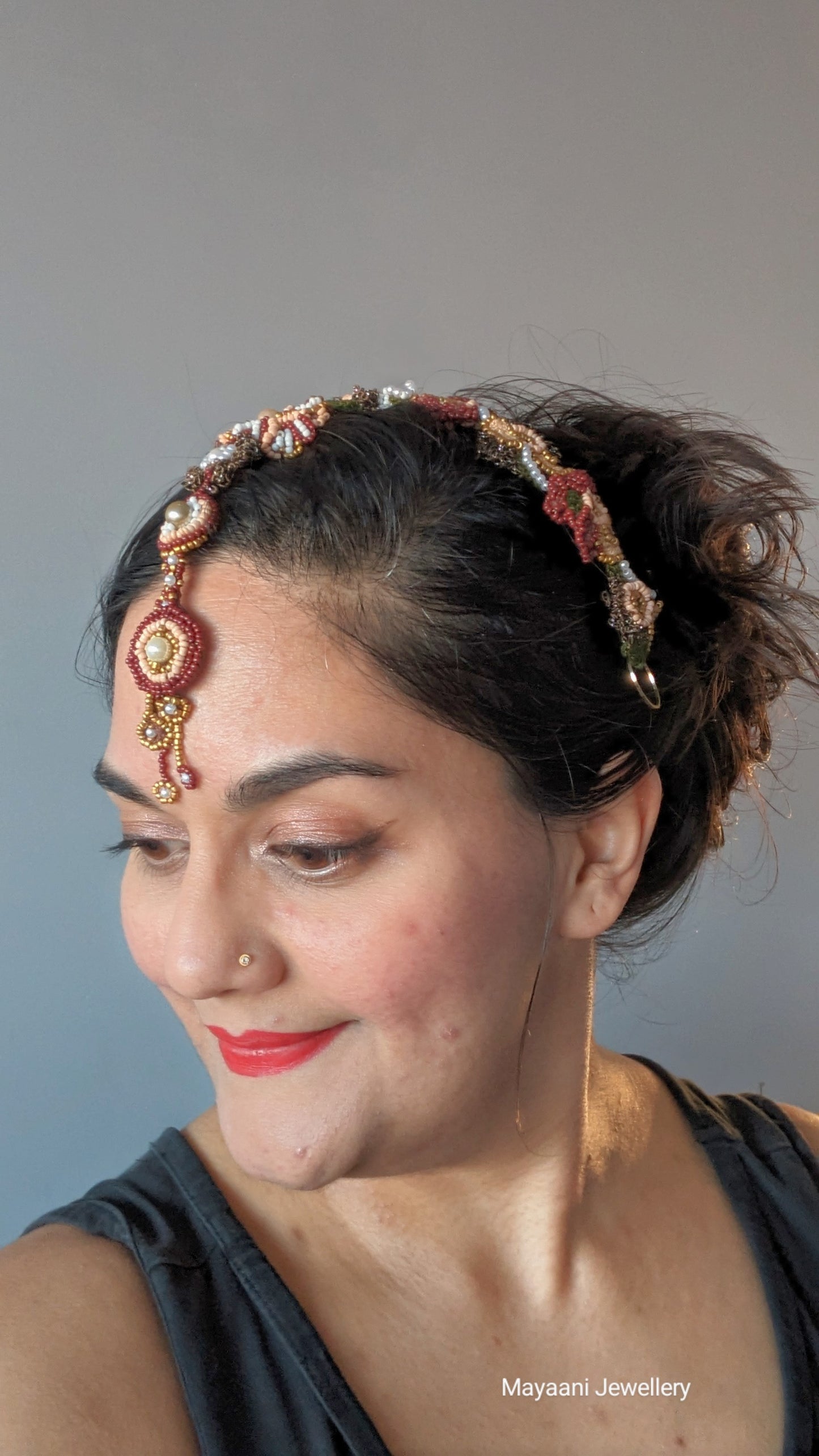 Enhance your style with this unique lightweight Indian bridal headpiece (maang tikka), showcased on a model for added inspiration. Designed to be worn as both a headband and tikka, this exclusive piece from Mayaani Jewellery UK blends traditional bridal elegance with modern sophistication. Hand-crocheted and beaded in vibrant deep brick red, gold, pearly white, and rich greens and browns, it reflects exquisite craftsmanship and artistic detail.