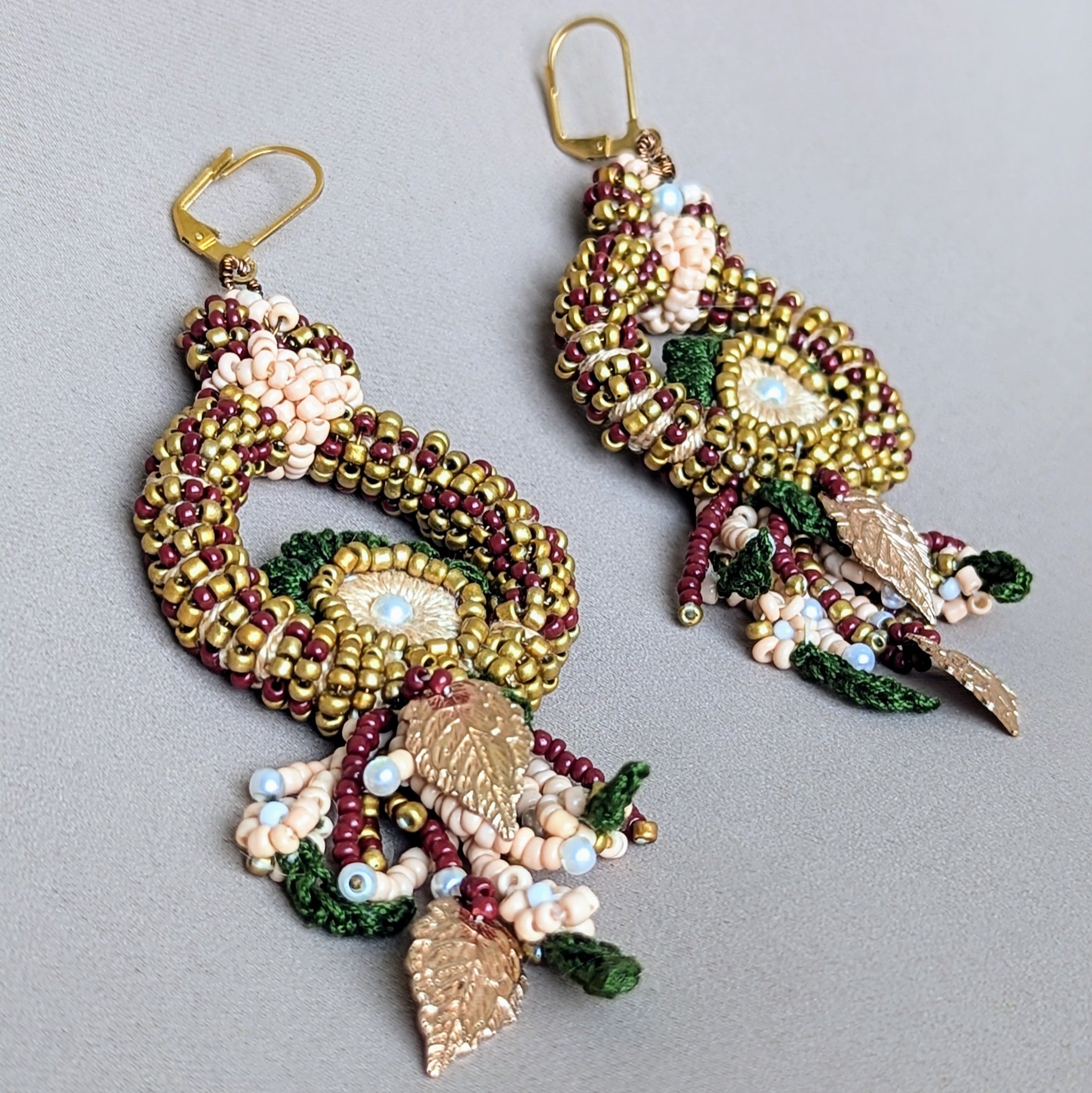 A unique one of a kind bridal pair of red green and gold bead crochet wire wrapped Indian earrings exclusive to sustainable UK brand Mayaani Jewellery. Featuring delicate floral and leaf like crochet elements adorned with intricate beadwork and accented with rose gold leaves for a peacock aesthetic perfect for weddings and special occasions.