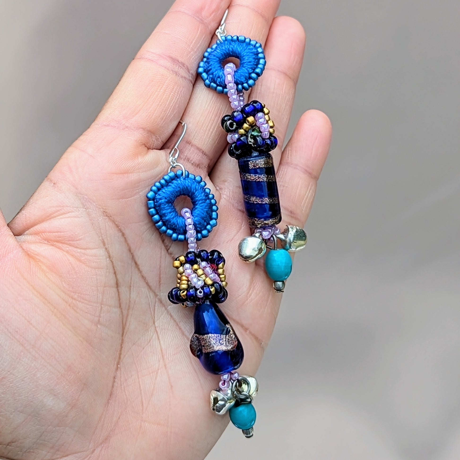 Hand holding a pair of Mayaani Jewellery UK earrings featuring beads uniquely made using scraps from in-house designer Megha's jewellery making process. The earrings feature a vibrant mix of blue, purple and turquoise beads, along with solid silver ghungroo bells which are synonymous with classical Indian dance for sustainable fashion steeped in culture.