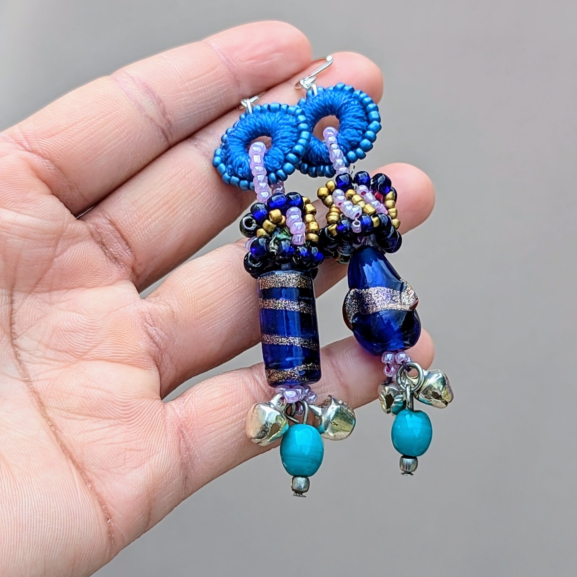 Hand holding a handcrafted pair of earrings by sustainable UK brand Mayaani Jewellery featuring unique scrap beads hand made from waste produced by in-house designer Megha's jewellery making process. Highlighted by rich blue and purple beads, hand crochet and turquoise accents, the earrings are finished with solid silver ghungroo bells, representing the spirit of classical Indian dance. A perfect balance of eco-friendly fashion and cultural craftsmanship.