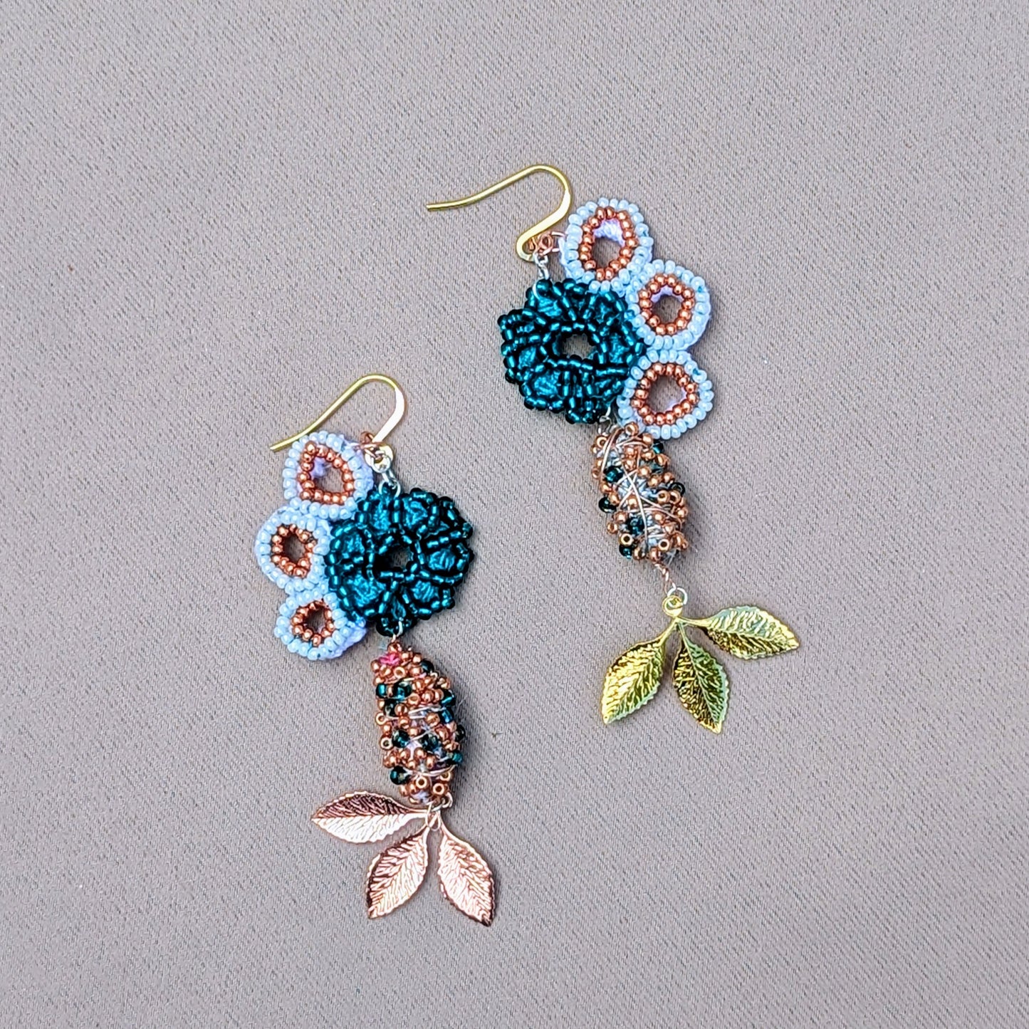 A pair of asymmetric earrings by Mayaani Jewellery featuring a seahorse motif, crafted from white and rose gold with turquoise accents. The design showcases hand-crocheted elements, copper details, and a vibrant mix of white and emerald green seed beads with teal hues. Gold and rose gold plated leaves cascade elegantly, highlighting organic shapes. Made with recycled materials, these sustainable earrings symbolize growth and renewal, perfect for weddings and special occasions.