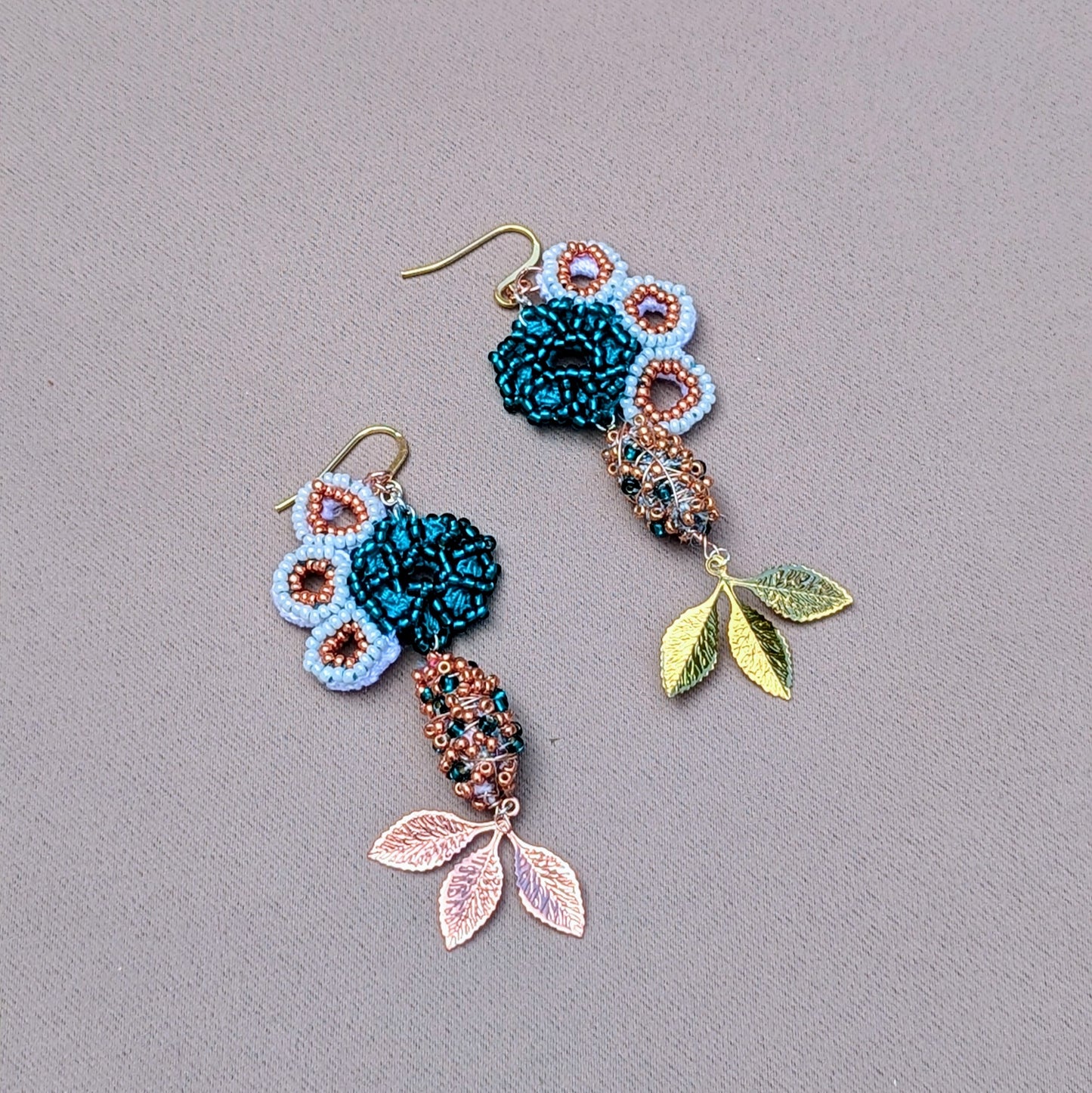 Unique asymmetric earrings by Mayaani Jewellery, designed with a seahorse motif. The earrings combine white and rose gold with turquoise, showcasing hand-crocheted details and copper elements. A colorful blend of white and emerald green seed beads, complemented by shimmering gold and rose gold plated leaves, evokes organic forms. Made from recycled materials, these earrings highlight sustainability and are perfect for special events, symbolizing growth and renewal.