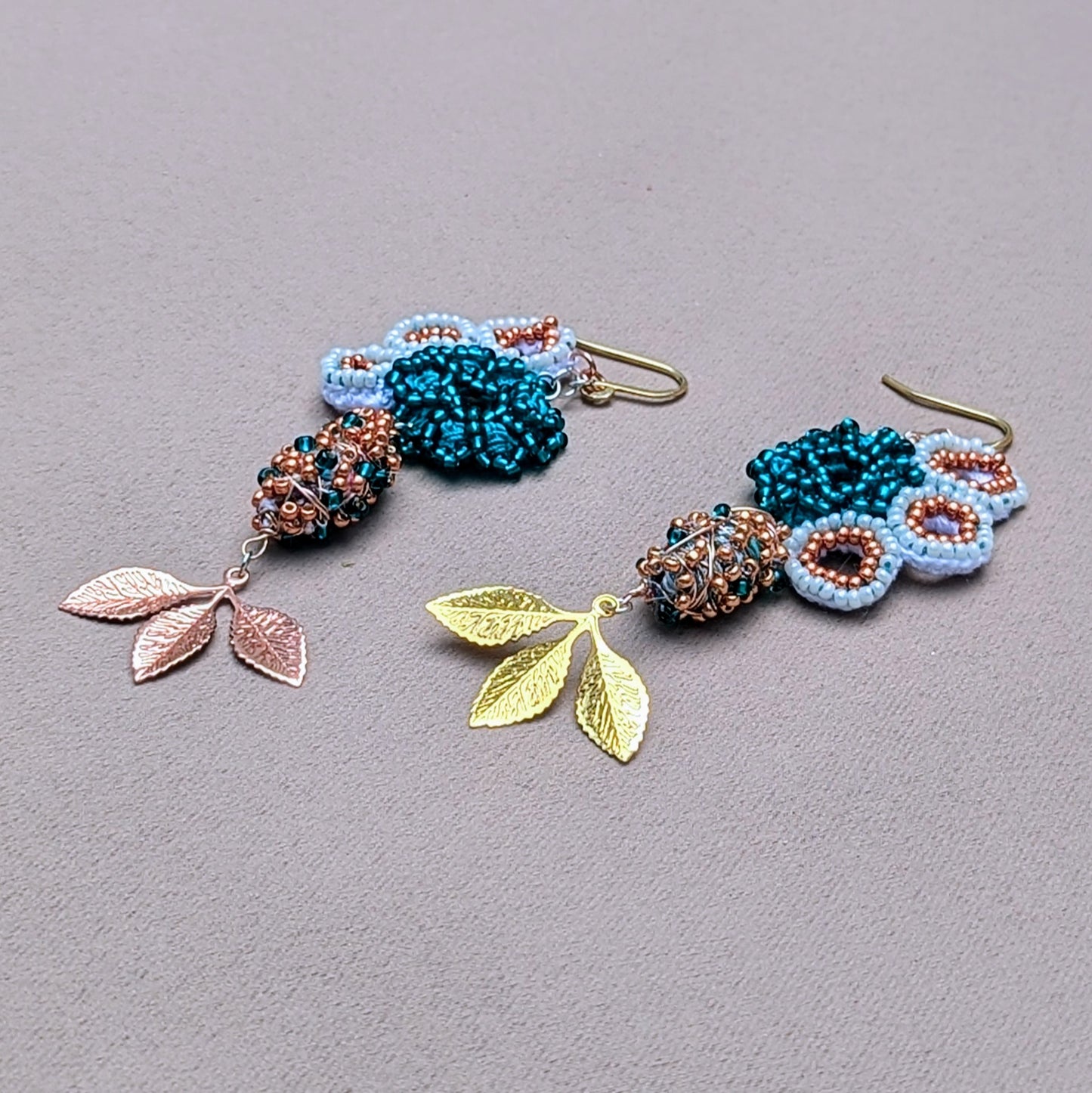 Asymmetric seahorse motif earrings by Mayaani Jewellery, combining white and rose gold with turquoise accents. The design includes hand-crocheted elements, copper accents, and a vibrant mix of white and emerald green seed beads, complemented by gold and rose gold plated leaves. Made from recycled materials, these sustainable earrings are perfect for weddings and special occasions, symbolizing growth and renewal through their organic-inspired design.