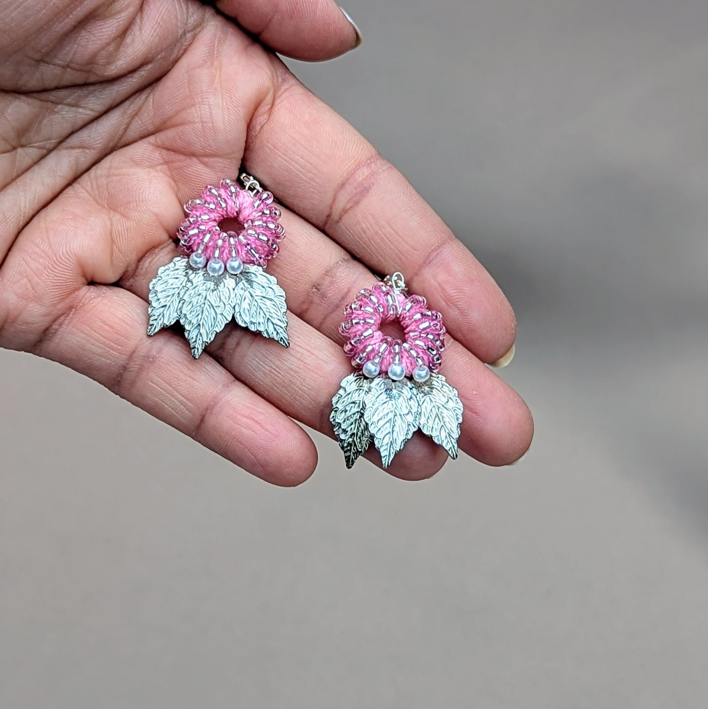 A unique pair of handcrafted earrings from the sustainable UK brand Mayaani Jewellery, featuring a pink beaded circle adorned with pearls, and intricated silver leaf designs dangling below. These earrings combine eco-conscious materials with a refined, nature-inspired aesthetic.