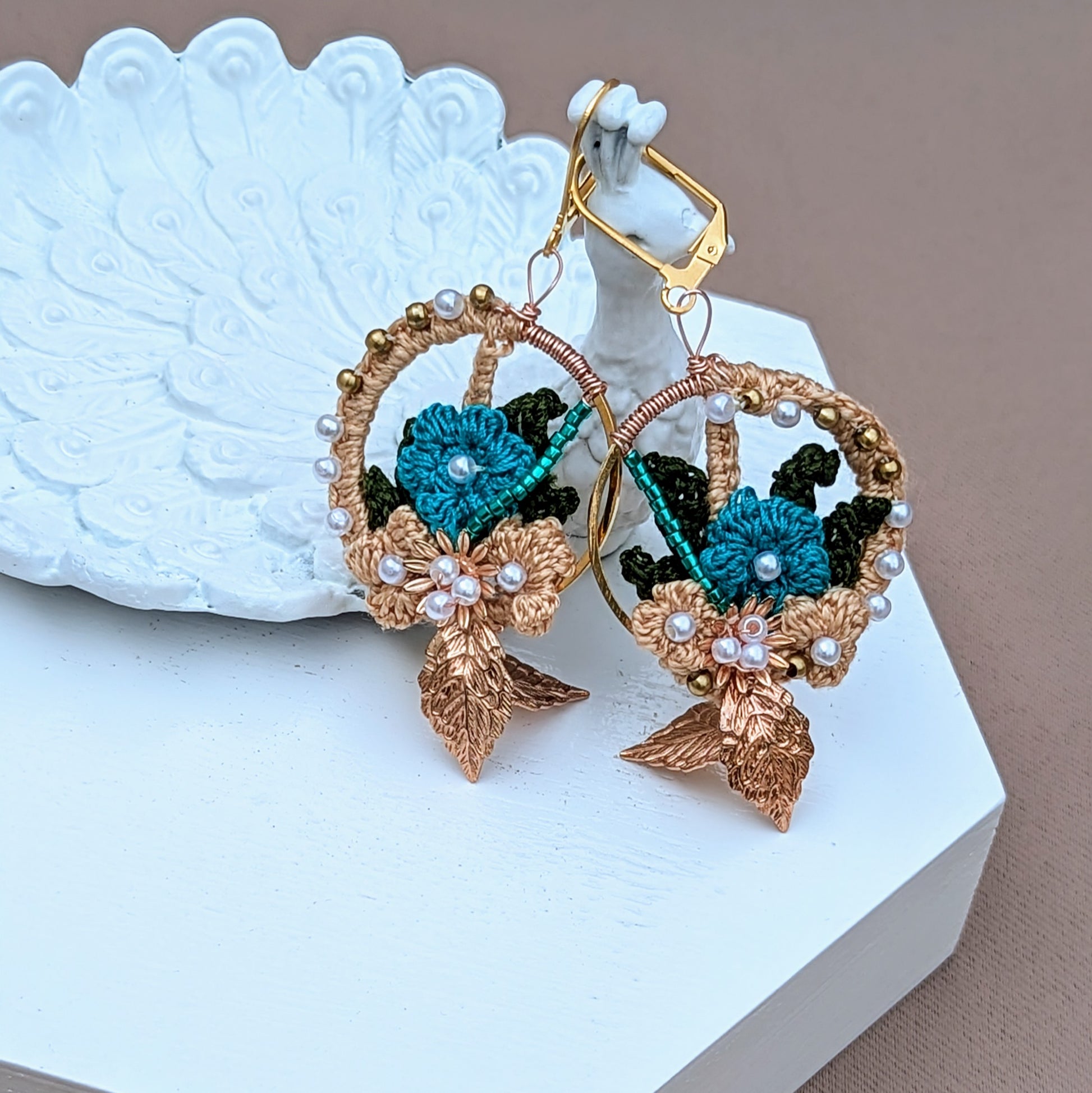 Pictured on a white peacock trinket dish, experience the elegance of these unique, handcrafted Indian-inspired earrings, exclusively from Mayaani Jewellery, a UK brand focused on sustainability. Designed with solid brass, seed pearls, and blue beads, and adorned with gold, blue, and dark green crochet floral motifs, these lightweight earrings are versatile enough for both special events and everyday wear.