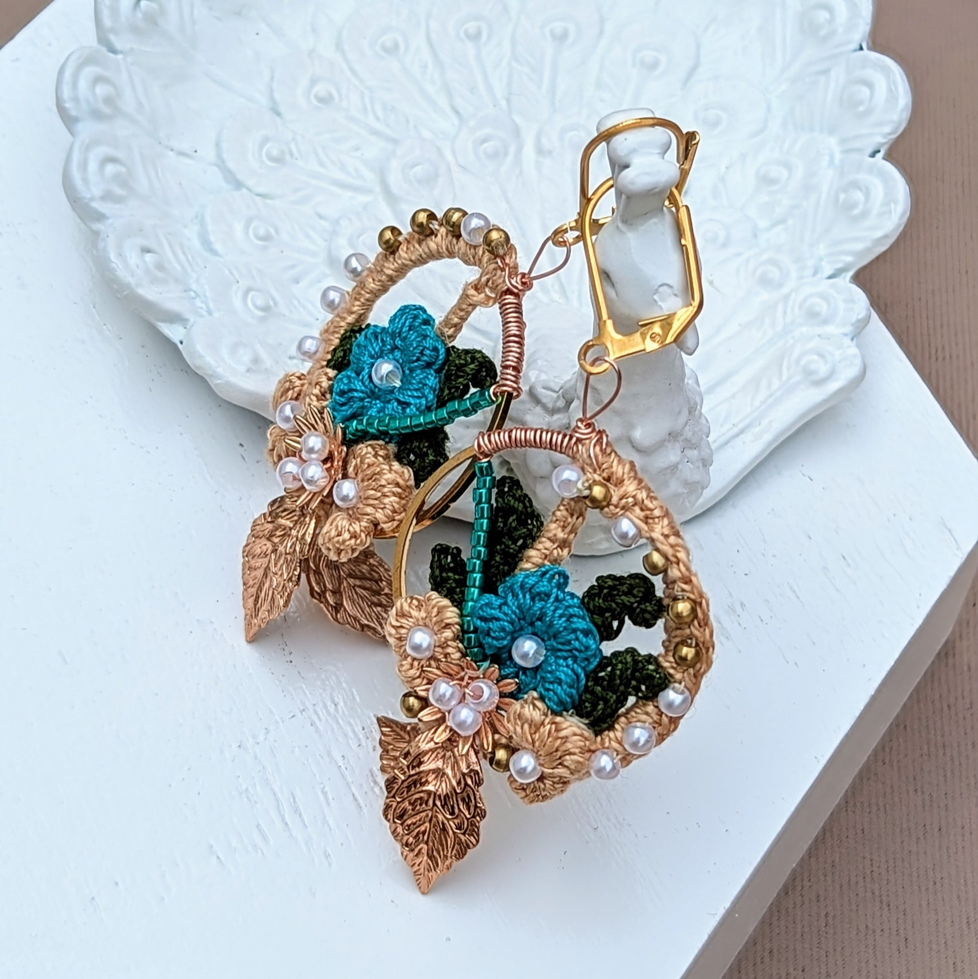 Embrace the artistry of these handcrafted Indian-inspired earrings, available exclusively from Mayaani Jewellery, a UK brand dedicated to sustainability. Pictured on a white peacock trinket dish, this pair feature intricate solid brass work, seed pearls, and blue bead accents with gold, blue, and dark green crochet floral patterns, these lightweight earrings offer both style and comfort for any occasion.