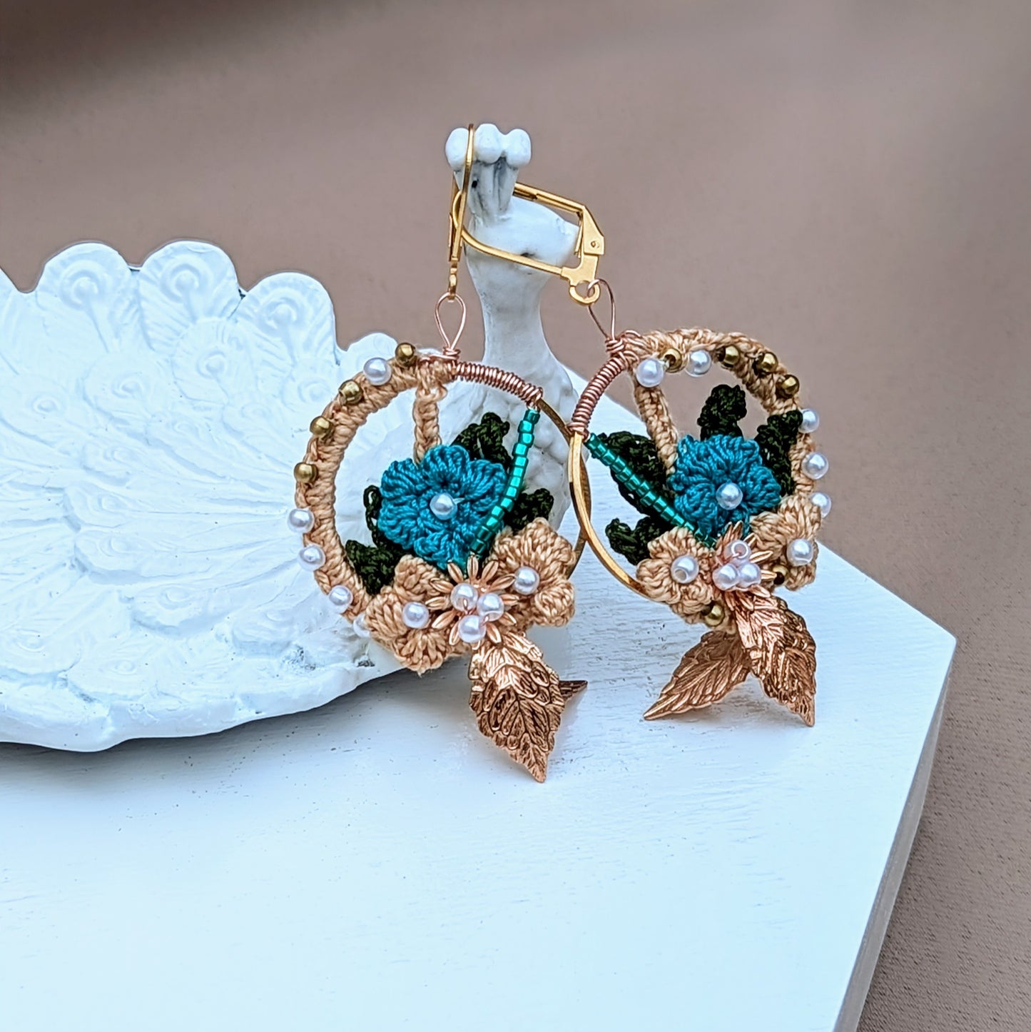 Elevate your accessory collection with these unique artisan-crafted Indian-inspired earrings from Mayaani Jewellery, a UK brand known for its sustainable practices. Pictured on a white peacock trinket dish they are adorned with intricately woven solid brass, seed pearls, and blue beads, and feature gold, blue, and dark green crochet floral motifs. These lightweight and comfortable earrings are suitable for both special occasions and daily wear.