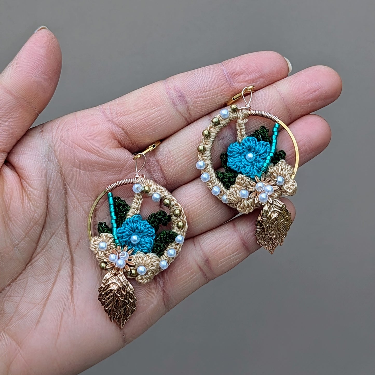 Explore the unique charm of these artisan-crafted Indian-inspired earrings, pictured in the palm of a hand and exclusively from Mayaani Jewellery, a sustainable UK brand. Artfully woven with solid brass, seed pearls, and blue beads, they showcase gold, blue, and dark green crochet floral designs. Lightweight and comfortable, these earrings are ideal for weddings, special events, or everyday elegance.