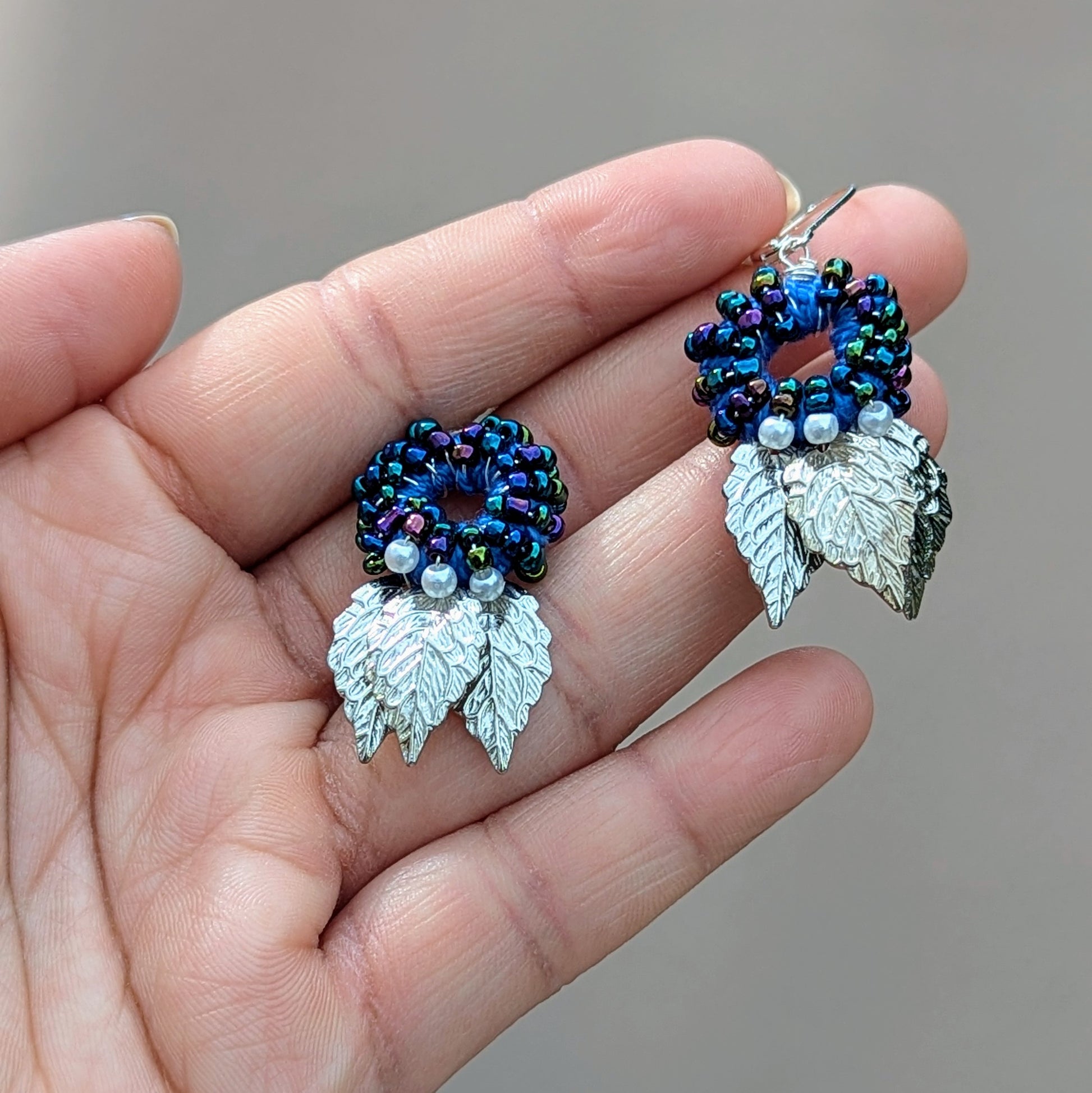 A hand holding a distinctive pair of artisan-crafted Indian-style earrings from the sustainable UK brand Mayaani Jewellery. These earrings boast an eye-catching circular blue bead design, adorned with elegant ivory seed pearls and silver leaf accents. Each piece is thoughtfully handmade, showcasing a luxurious mix of hand crochet, intricate beadwork, and skilled wire wrapping, finished with 925 stamped lever-back hooks for a chic and contemporary look that honours traditional craftsmanship.