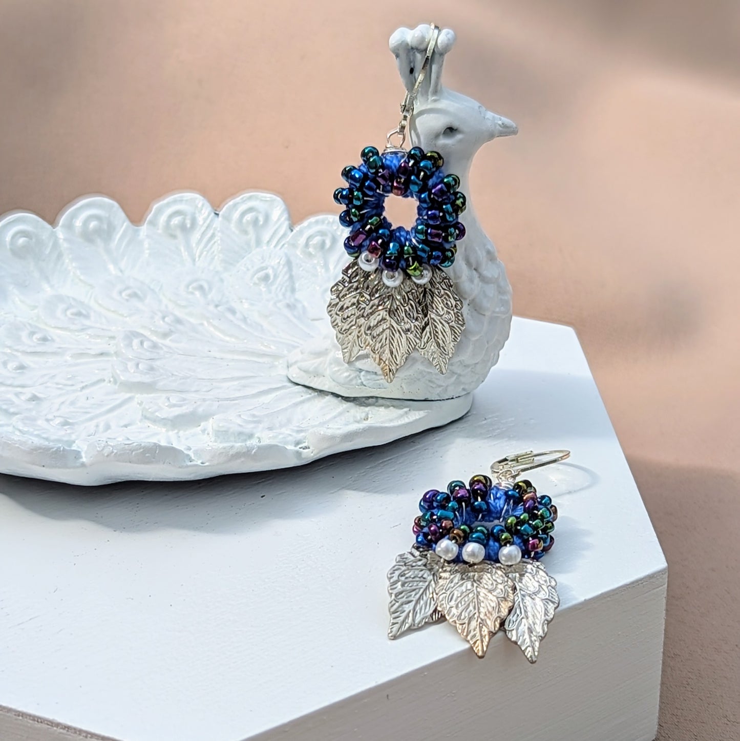 Presenting a unique pair of artisan-crafted Indian-style earrings by the sustainable UK brand Mayaani Jewellery displayed on a white peacock. These earrings feature a captivating circular blue bead design, enhanced by charming ivory seed pearls and silver leaf accents. Meticulously handmade, they showcase an exquisite combination of hand crochet, detailed beadwork, and expert wire wrapping, all beautifully completed with 925 stamped lever-back hooks for a stylish blend of modern and traditional artistry.