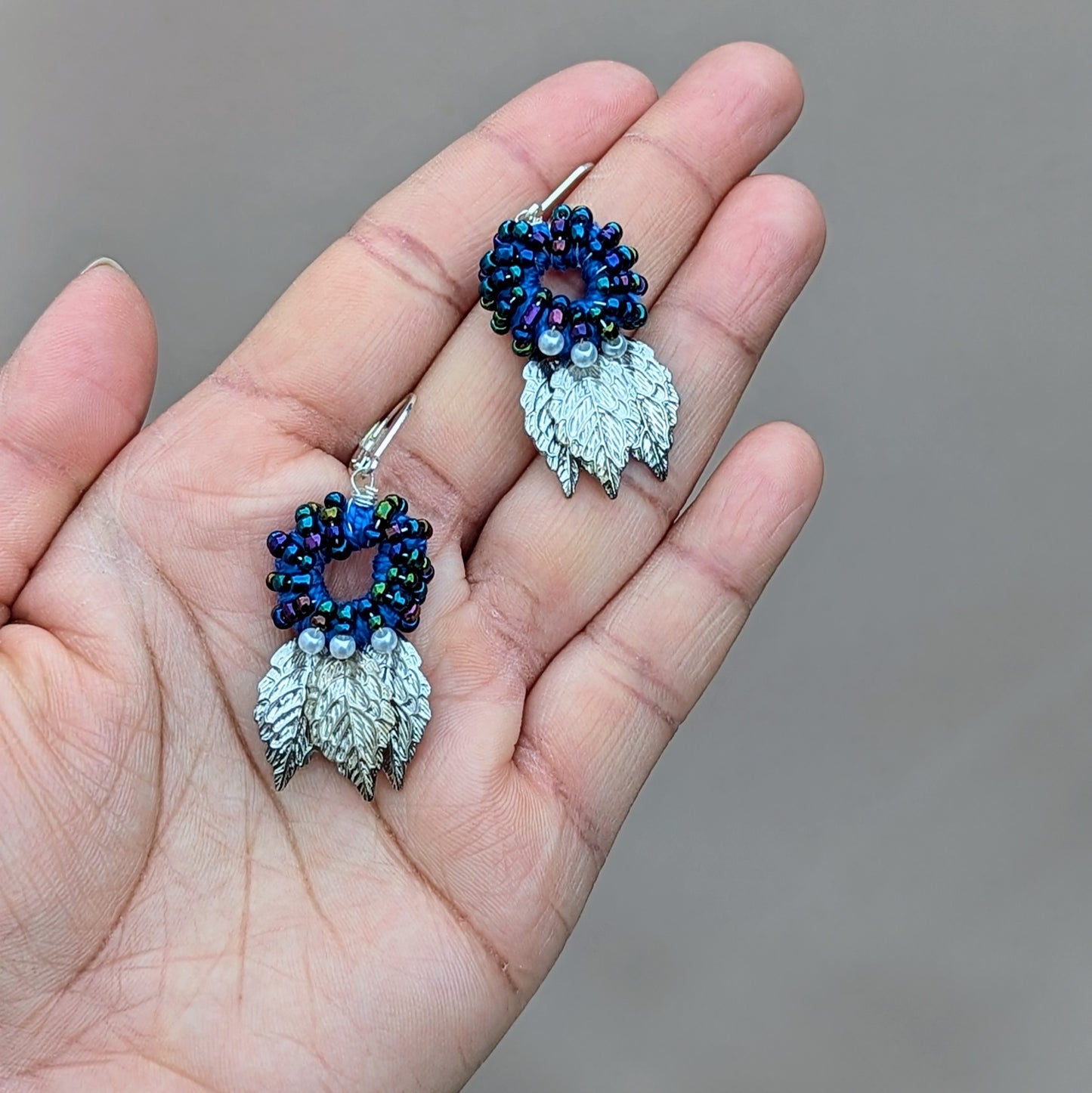 Introducing a stunning pair of artisan-crafted Indian-style earrings from the sustainable UK brand Mayaani Jewellery. These earrings highlight a beautiful circular blue bead design, complemented by delicate ivory seed pearls and silver leaf accents. Each piece is a testament to skilled craftsmanship, featuring a rich blend of hand crochet, meticulous beadwork, and precise wire wrapping, all elegantly finished with 925 stamped lever-back hooks for a contemporary yet timeless look.