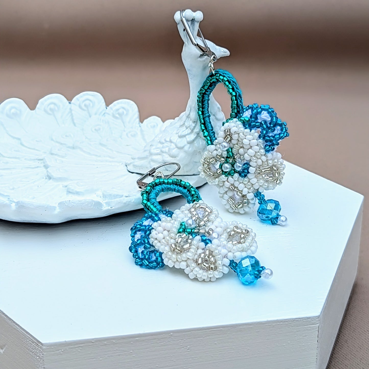 Discover a breath taking pair of handcrafted bridal earrings from the sustainable UK brand Mayaani Jewellery displayed on a peacock trinket dish. Featuring intricate hand crochet and floral designs, these earrings are finished with sleek stainless steel lever back clasps and high-quality upcycled Swarovski-style bicone beads, making them an ideal "something borrowed" for your wedding day.