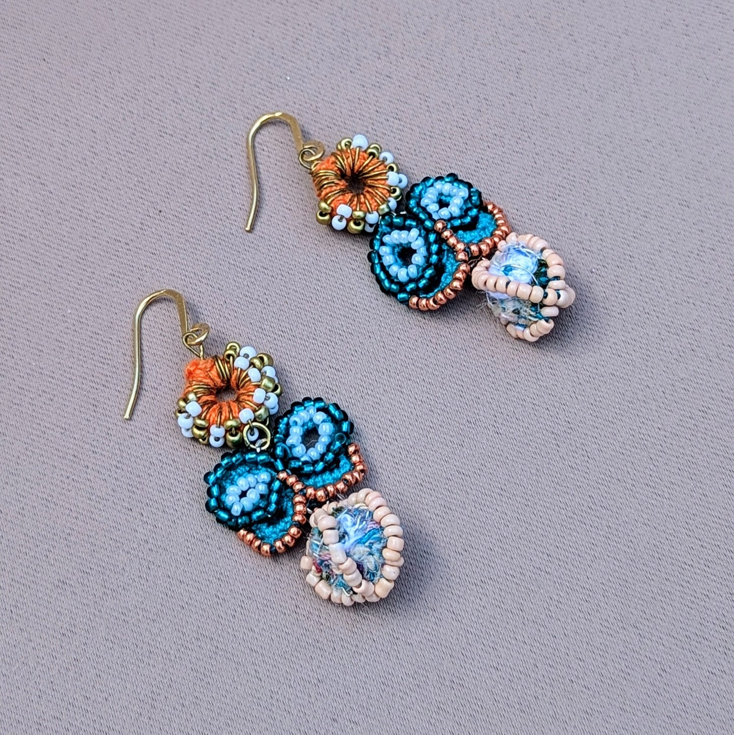 A pair of eco-friendly blue butterfly earrings handcrafted by sustainable brand Mayaani Jewellery UK. The earrings feature a turquoise and rose gold seed bead butterfly, set atop an orange crochet circle adorned with white and gold seed beads. The design includes a stunning scrap bead at the base, with durable gold-plated stainless steel hooks. Enjoy artistic Indian inspired style.