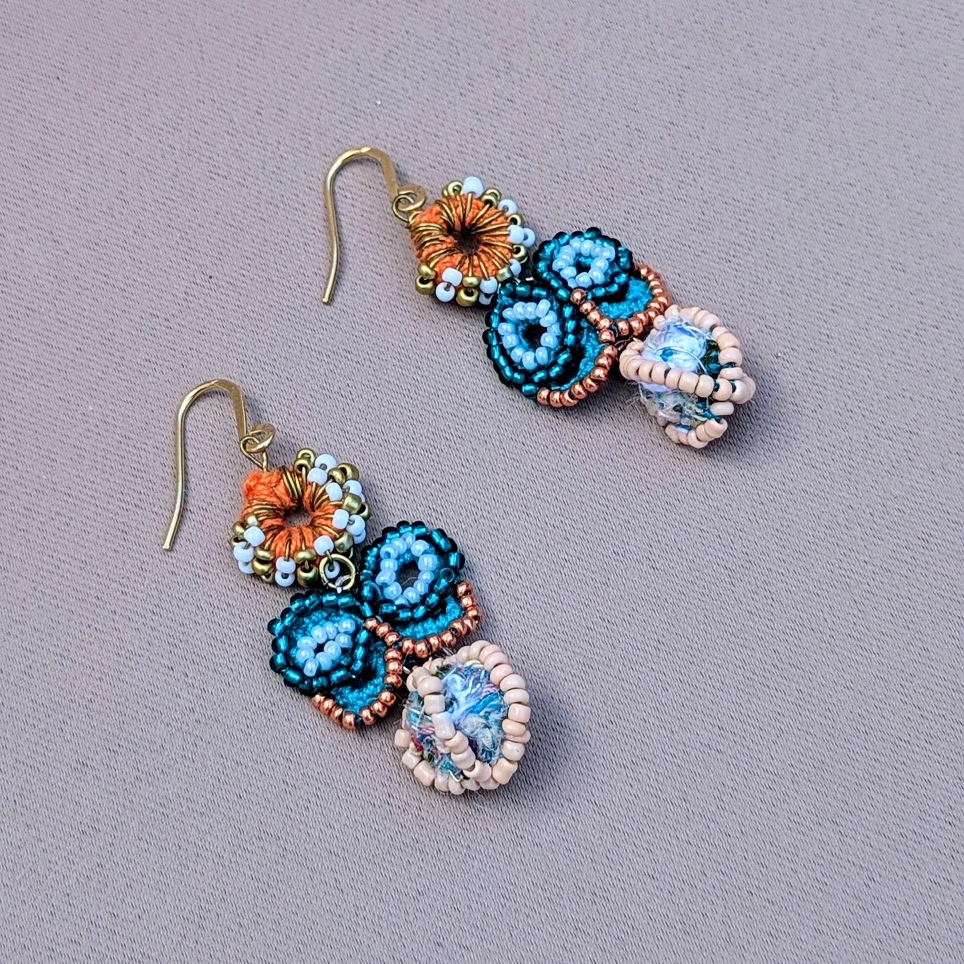 A pair of eco-friendly blue butterfly earrings handcrafted by sustainable brand Mayaani Jewellery UK. The earrings feature a turquoise and rose gold seed bead butterfly, set atop an orange crochet circle adorned with white and gold seed beads. The design includes a stunning scrap bead at the base, with durable gold-plated stainless steel hooks. Enjoy artistic Indian inspired style.