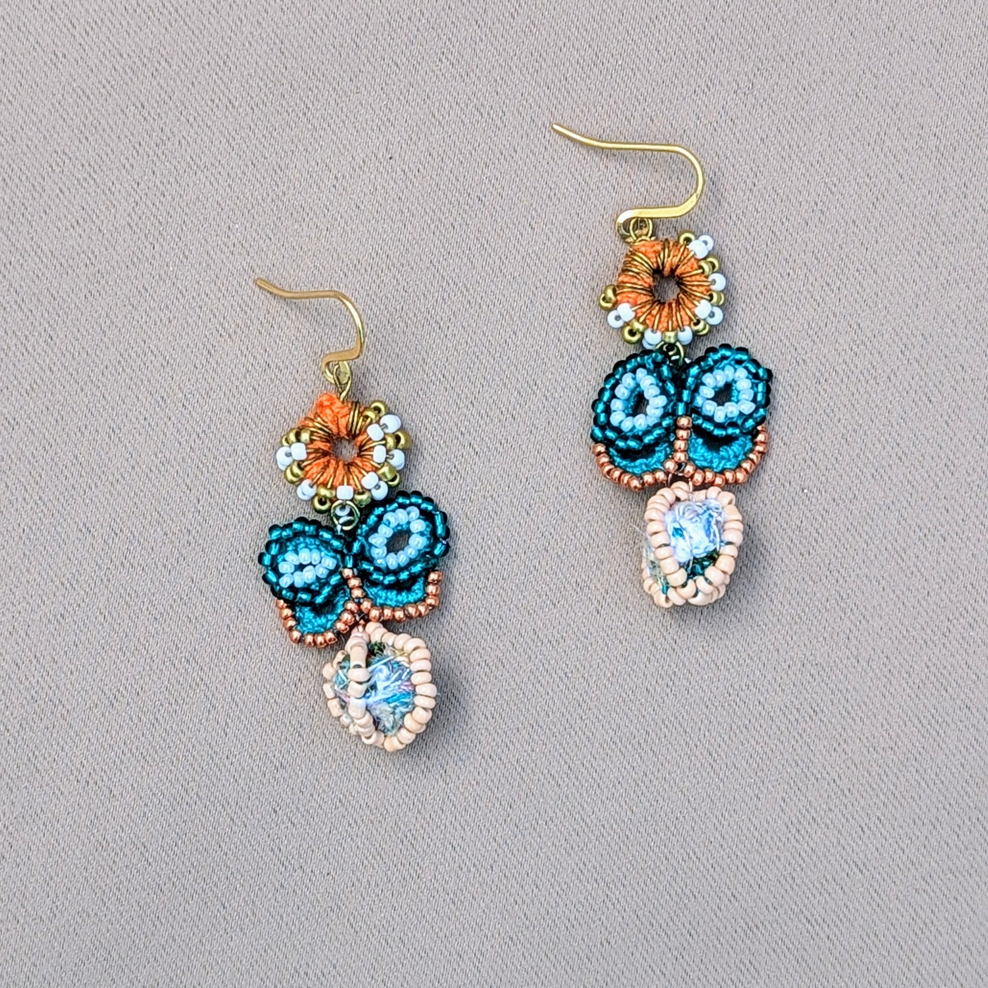 Handcrafted blue butterfly earrings from Mayaani Jewellery UK, showcasing a unique scrap bead as a focal point beneath a butterfly crafted from turquoise and rose gold seed beads. The earrings include an orange crochet circle with white and gold seed beads, secured by gold-plated stainless steel hooks.