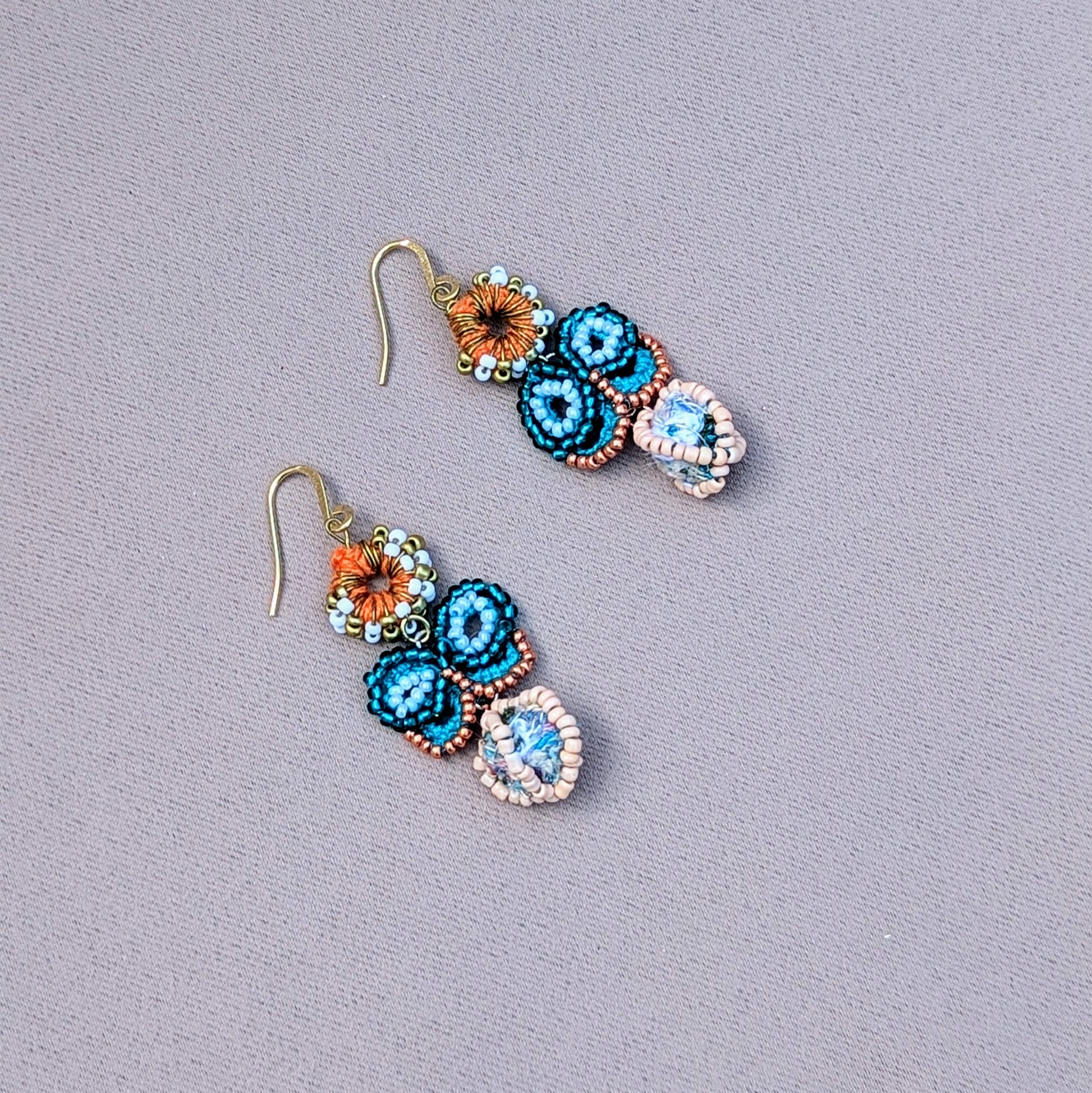 Sustainable blue butterfly earrings by Mayaani Jewellery UK, highlighting an enchanting scrap bead at the base of a turquoise and rose gold seed bead butterfly. The design is completed with an orange crochet circle embellished with white and gold seed beads and durable gold-plated stainless steel hooks. Experience the allure of cutting edge jewellery design drawing on Indian cultural influences.