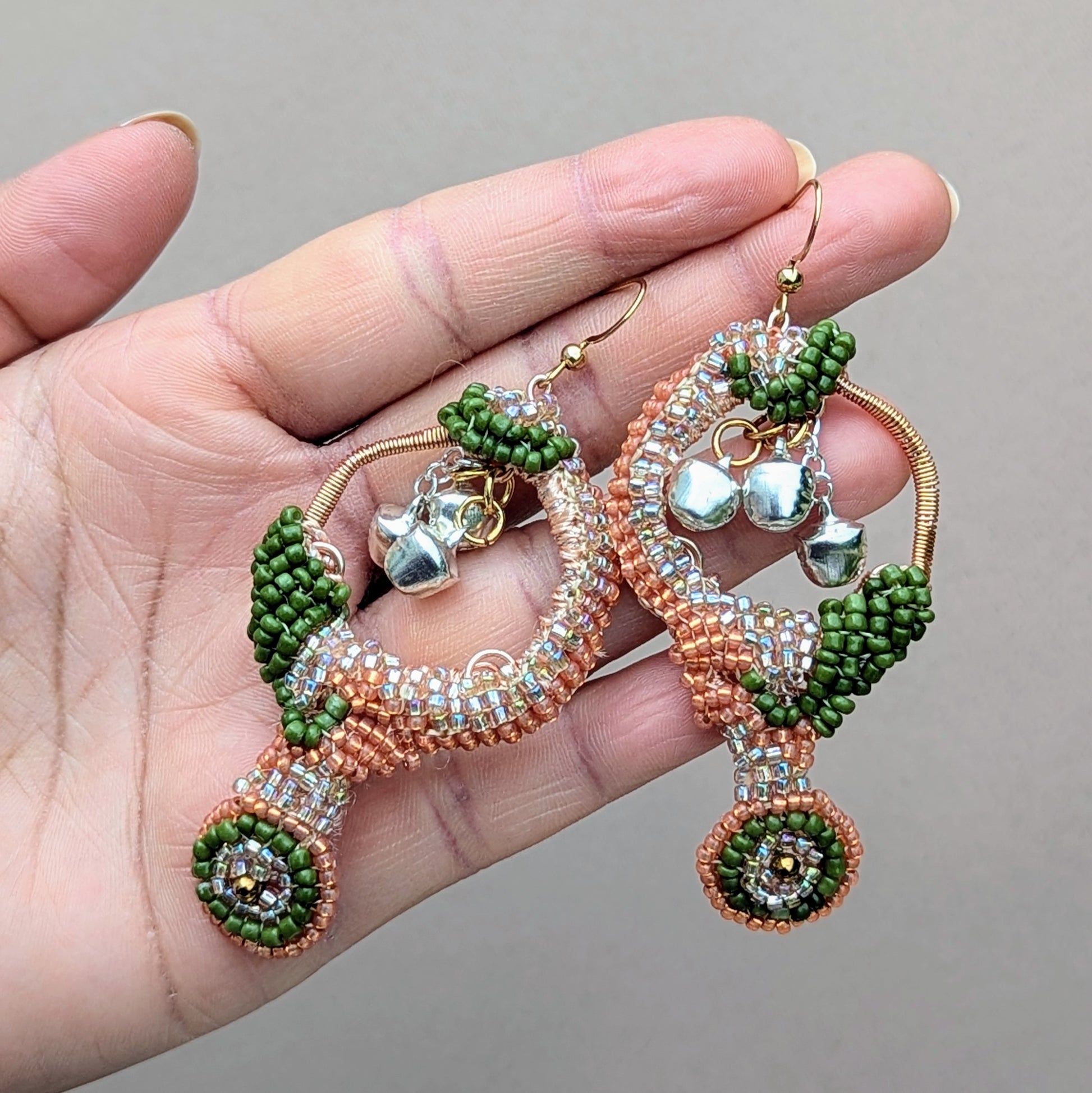 A hand displays a pair of lightweight, Indian-inspired earrings from the sustainable UK brand Mayaani Jewellery, worthy of a goddess. Designed to ignite the dancer within, these elegant earrings feature solid silver ghungroo bells that chime with each movement, celebrating your classical Indian dance heritage. Adorned with luxurious silver, peach, and green beads, along with hand crochet and wire wrapping, they are the perfect dazzling accessory for weddings and special events.