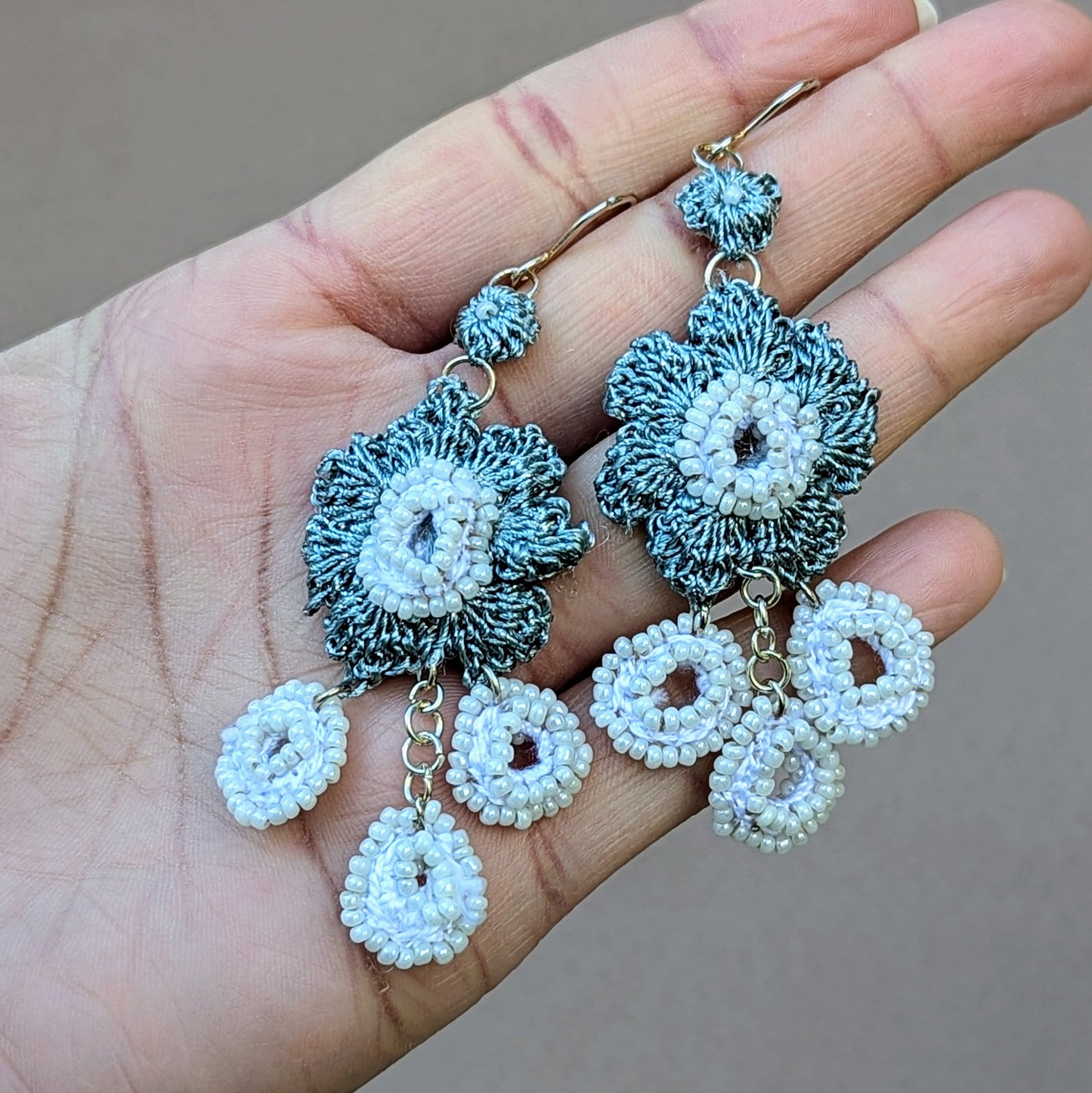 Discover a unique, one-of-a-kind pair of artisan handcrafted dangle drop earrings from Mayaani Jewellery, a sustainable UK brand. Inspired by Indian design, these earrings feature beaded circular motifs that elegantly dangle from a bold central floral accent. Crafted with a rich blend of white, metallic grey hand crochet, white seed beads, and silver jump rings, they are the perfect statement accessory for bridal, wedding, and special occasions. Complete the look with a matching hairpin and a hand bracelet.