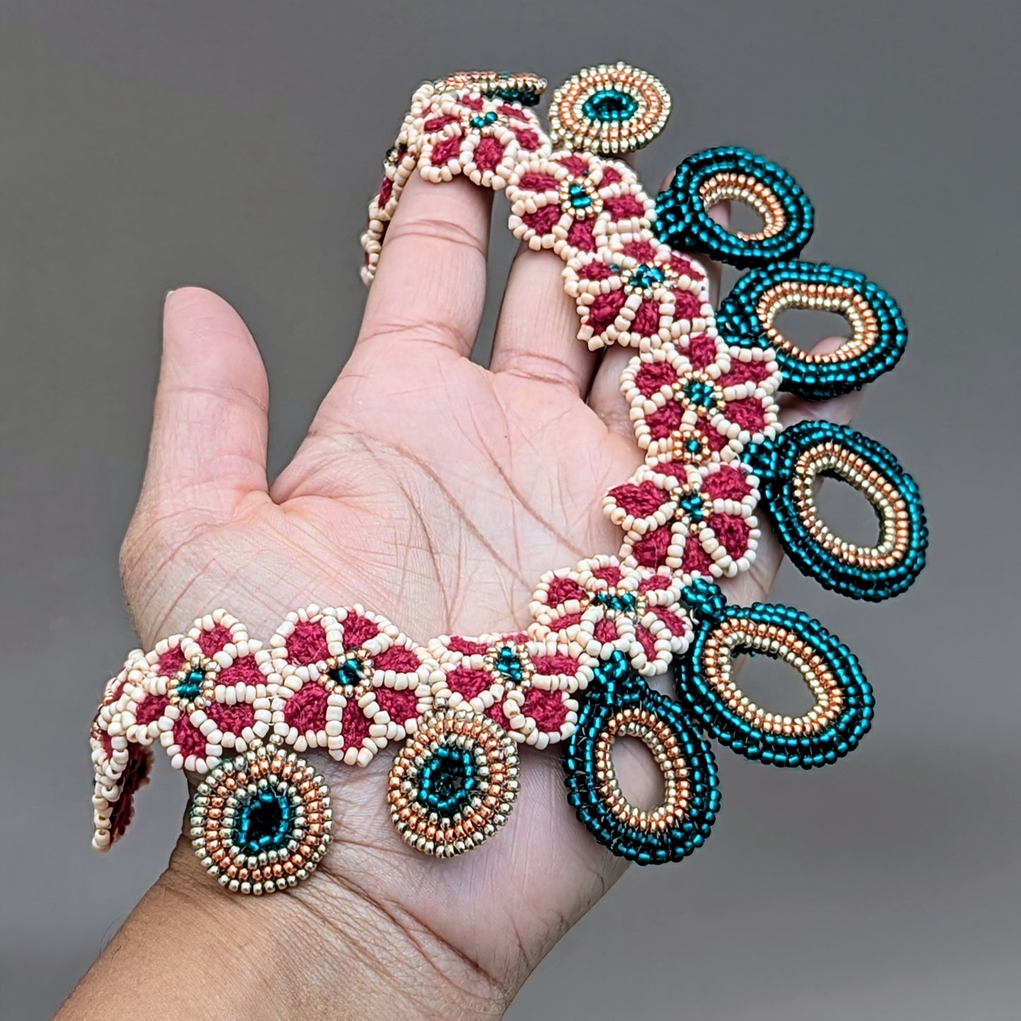 Hand holding a unique handcrafted necklace from sustainable UK brand Mayaani Jewellery inspired by Indian tradition. This piece features detailed hand crochet, beadwork and wirework in red, emerald green, and peach tones. Accented with rose gold and gold, it reimagines traditional Indian bridal colours with a contemporary flair. The necklace combines floral motifs with circular bead pendants, creating an eye-catching and elegant design.