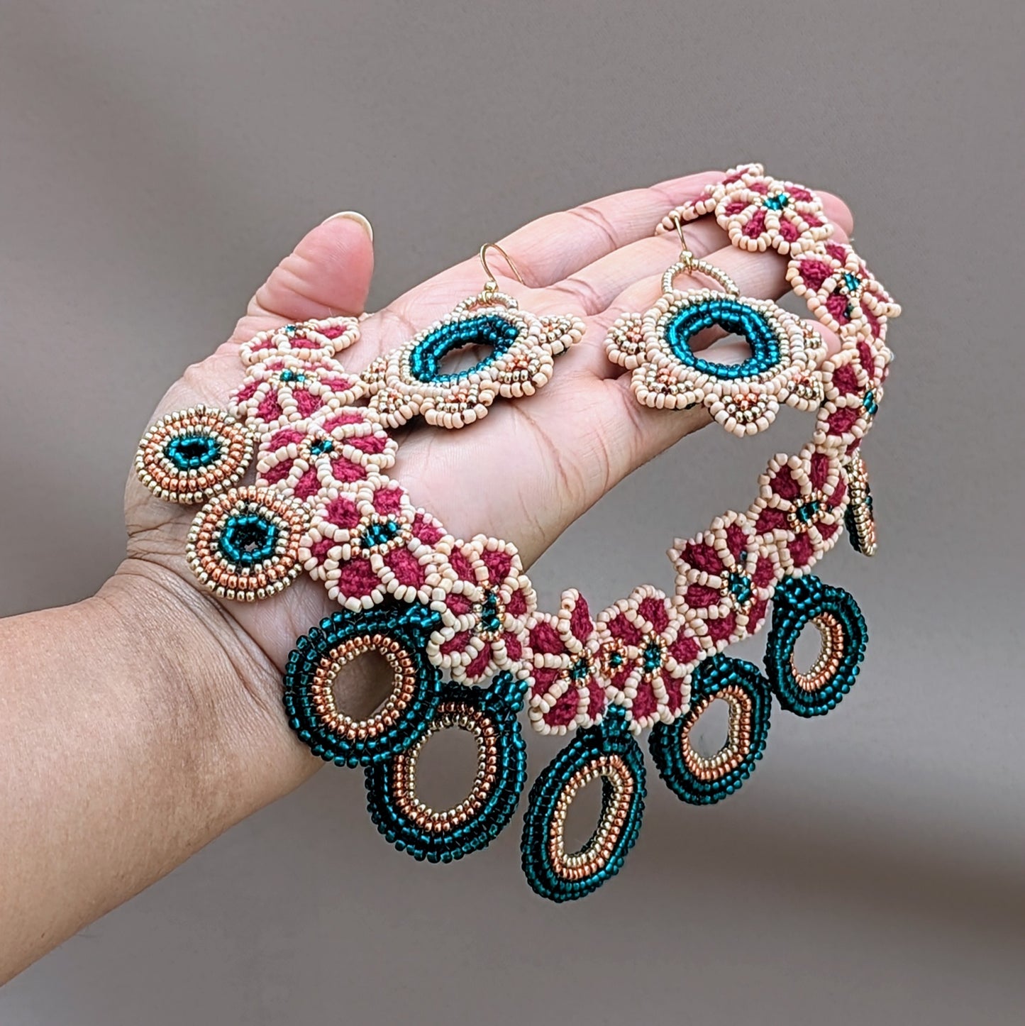 Hand holding a one-of-a-kind handcrafted necklace from UK-based Indian wedding & occasion brand Mayaani Jewellery, combining Indian bridal inspiration with modern design. Featuring delicate hand crochet, beadwork, and wirework in vibrant red, emerald green, and peach, it is accentuated by rose gold and gold details. The necklace showcases floral motifs alongside bold circular beaded pendants, creating an exquisite fusion of traditional and contemporary styles.