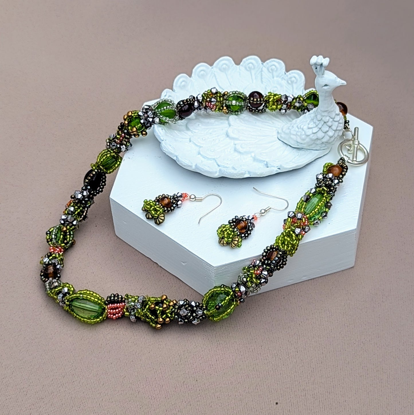 Embrace sustainable style with this artisan handcrafted beaded choker necklace, displayed on a white display stand, from Mayaani Jewellery, a sustainable Indian UK brand. Celebrate the beauty of nature's seasons through a vibrant blend of greens and browns, featuring new and high-quality upcycled glass seed beads. Expertly woven and artistically intertwined, this statement piece is perfect for any outfit and comes with a matching pair of short drop earrings.