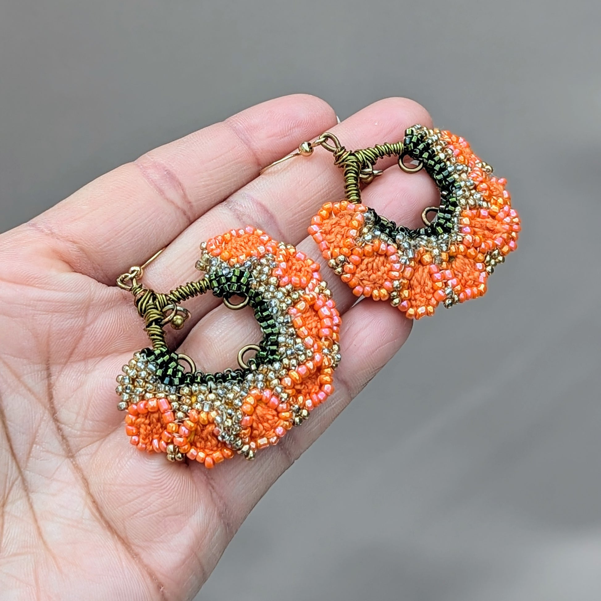 Introducing a striking pair of Indian inspired orange, green, and gold earrings, exclusively from the sustainable UK brand Mayaani Jewellery. These earrings showcase a charming brass bead that gracefully dangles from intricately wrapped bronze copper wire, adding delightful movement to the floral fan-like design. Perfect for both special occasions and everyday wear, they bring pure joy to any look.