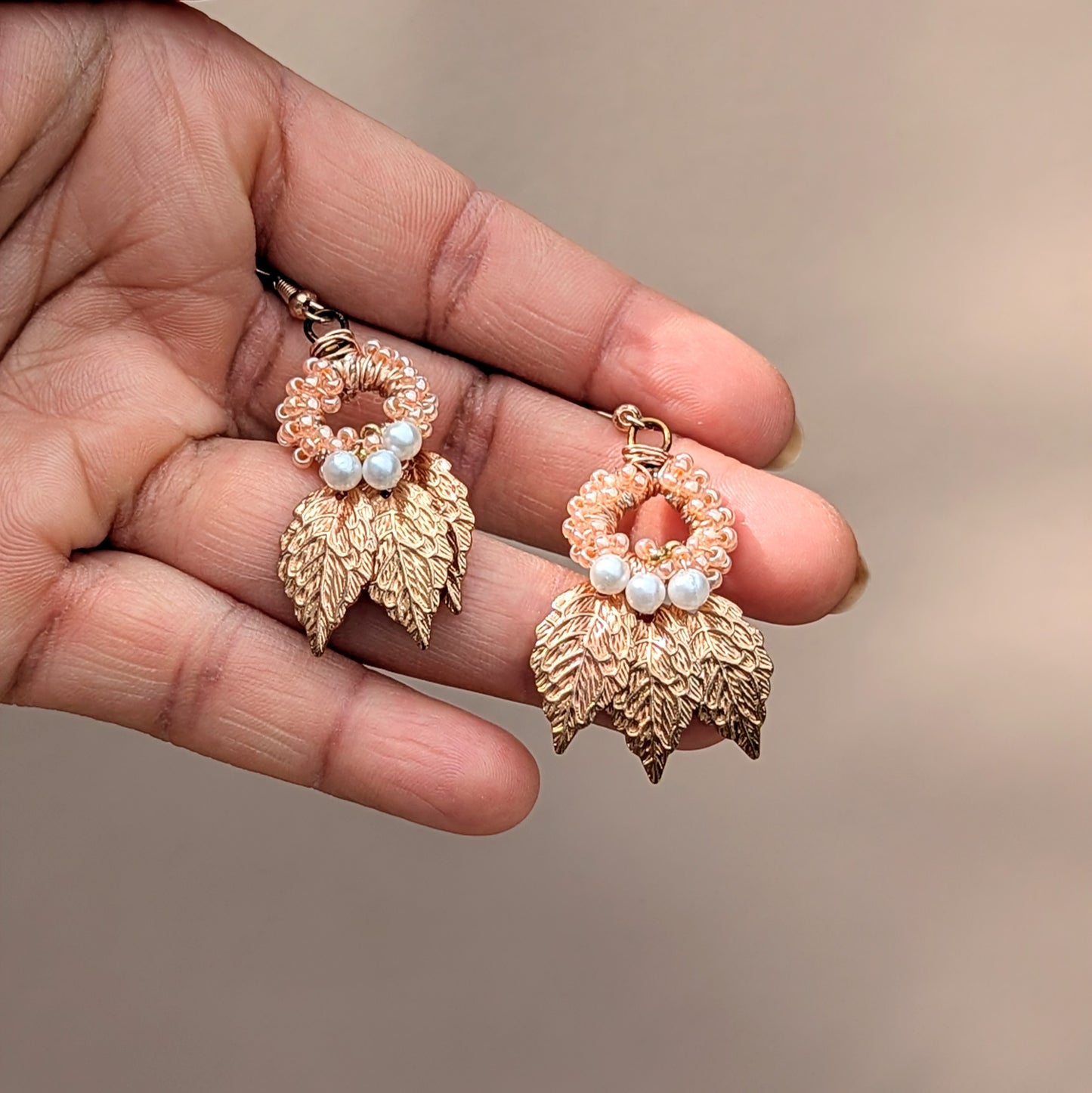 Indian Boho-inspired earrings in the palm of a hand exclusive to sustainable UK brand Mayaani Jewellery. Crafted with gold-plated leaf pendants and adorned with hand-crocheted peach beadwork and seed pearls. These artisan earrings feature a blend of hand wire work and are one colour combination of a limited edition best selling design from the UK-based brand focussed on eco-conscious fashion.