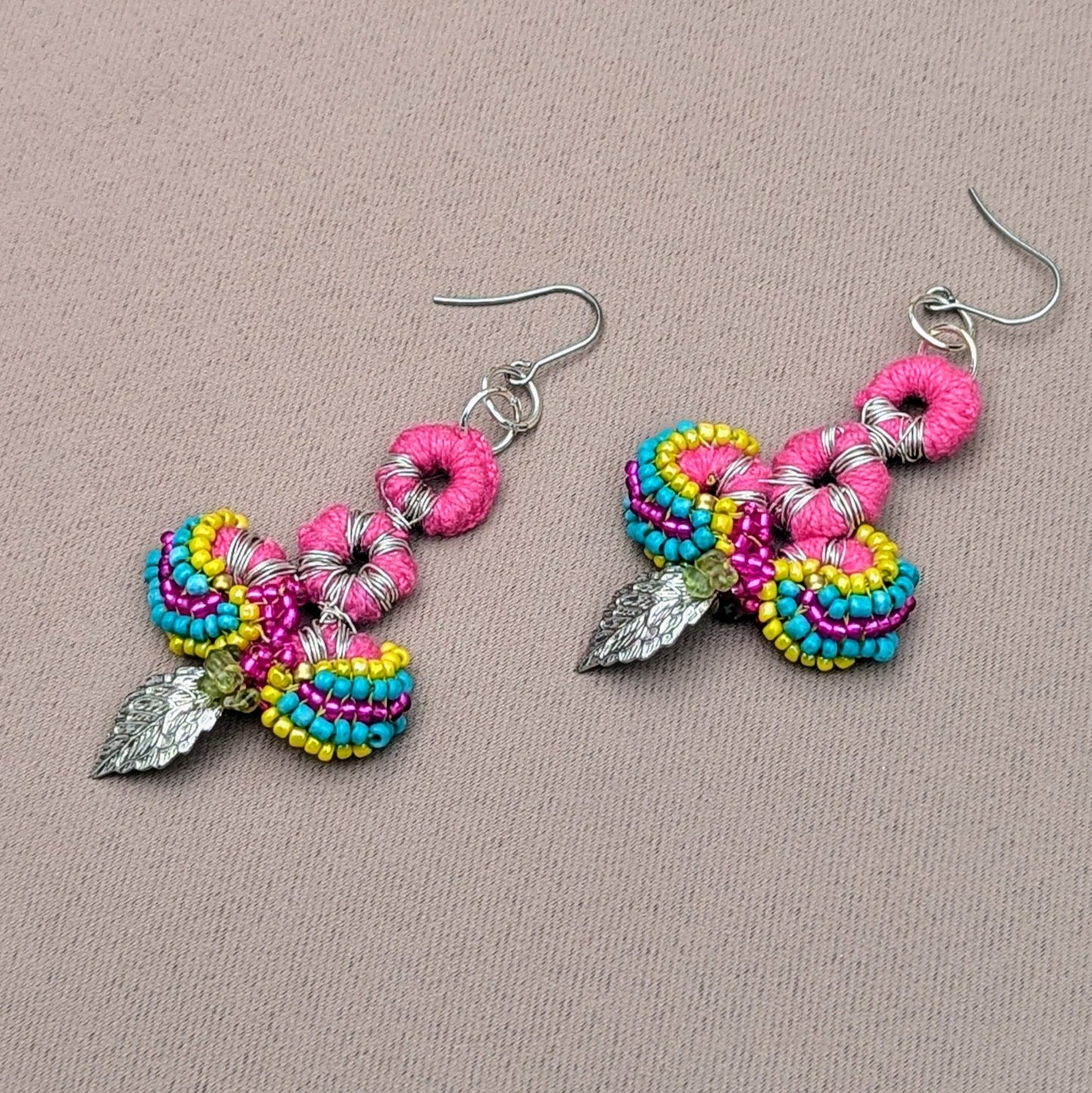 A vibrant pair of handcrafted earrings by Mayaani Jewellery, a sustainable UK brand known for its Indian-inspired designs. These earrings feature intricate pink crochet work, colourful bead patterns in turquoise, yellow, and purple, and elegant silver leaf charms. Made using high-quality upcycled materials, including peridot gemstone chips, they are exceptionally lightweight and comfortable, showcasing Mayaani's dedication to eco-friendly craftsmanship and artistic detail.