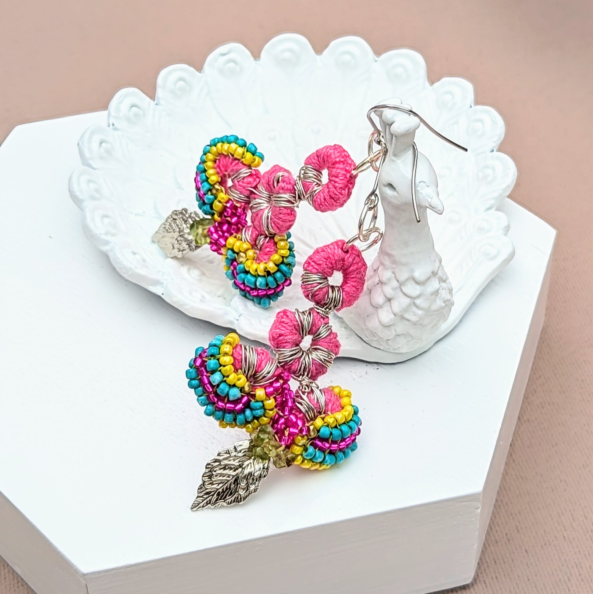 Displayed on a white peacock trinket dish and handcrafted by the sustainable UK brand Mayaani Jewellery, these Indian-inspired earrings feature a vibrant mix of colours with intricate pink crochet, multi-coloured beadwork, and silver leaf charms. Made from upcycled materials, including peridot chips, they are lightweight  and eco-conscious. Their bold yet delicate design embodies Mayaani's focus on sustainability, artistry and high-quality craftsmanship.