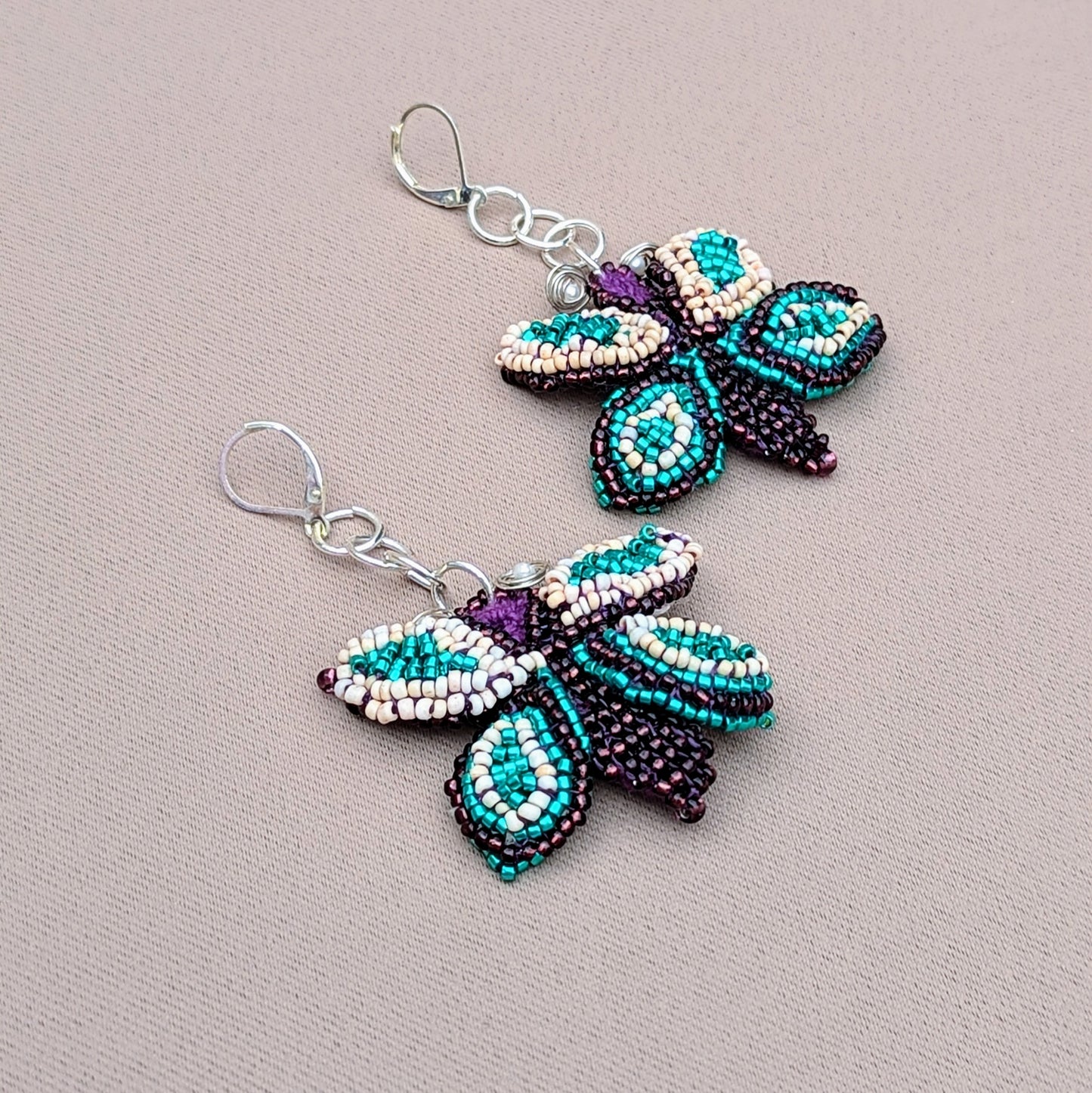 These exclusive butterfly statement earrings from Mayaani Jewellery, a sustainable UK brand, are artisan-crafted with exceptional beadwork on a hand-crocheted background. Detailed wire wrapping accents and 925 stamped sterling silver lever-back hooks complete the look. The harmonious blend of purple, soft creamy peach, and bold turquoise makes these earrings a statement piece for any outfit.