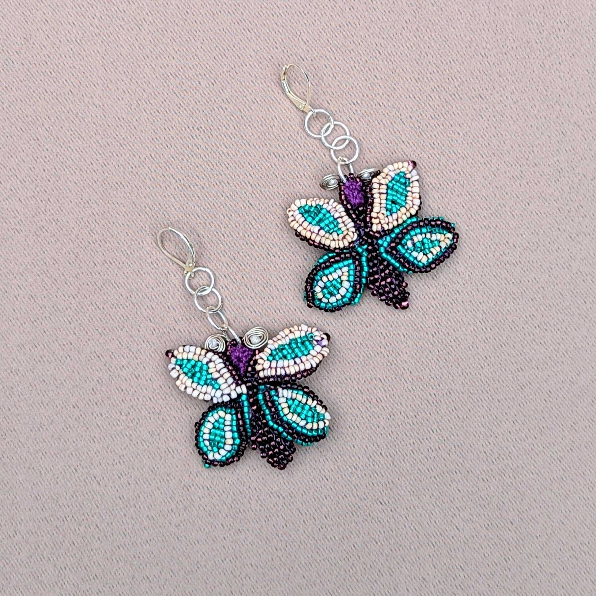 An exclusive pair of artisan handcrafted butterfly statement earrings from sustainable UK brand Mayaani Jewellery, showcasing exceptionally intricate beadwork on a hand-crocheted backdrop, accented with delicate hand wire wrapping. Finished with 925 stamped sterling silver lever-back hooks, these earrings are a stunning blend of purple, soft creamy peach, and vibrant turquoise, making them a bold statement piece for any occasion including everyday.