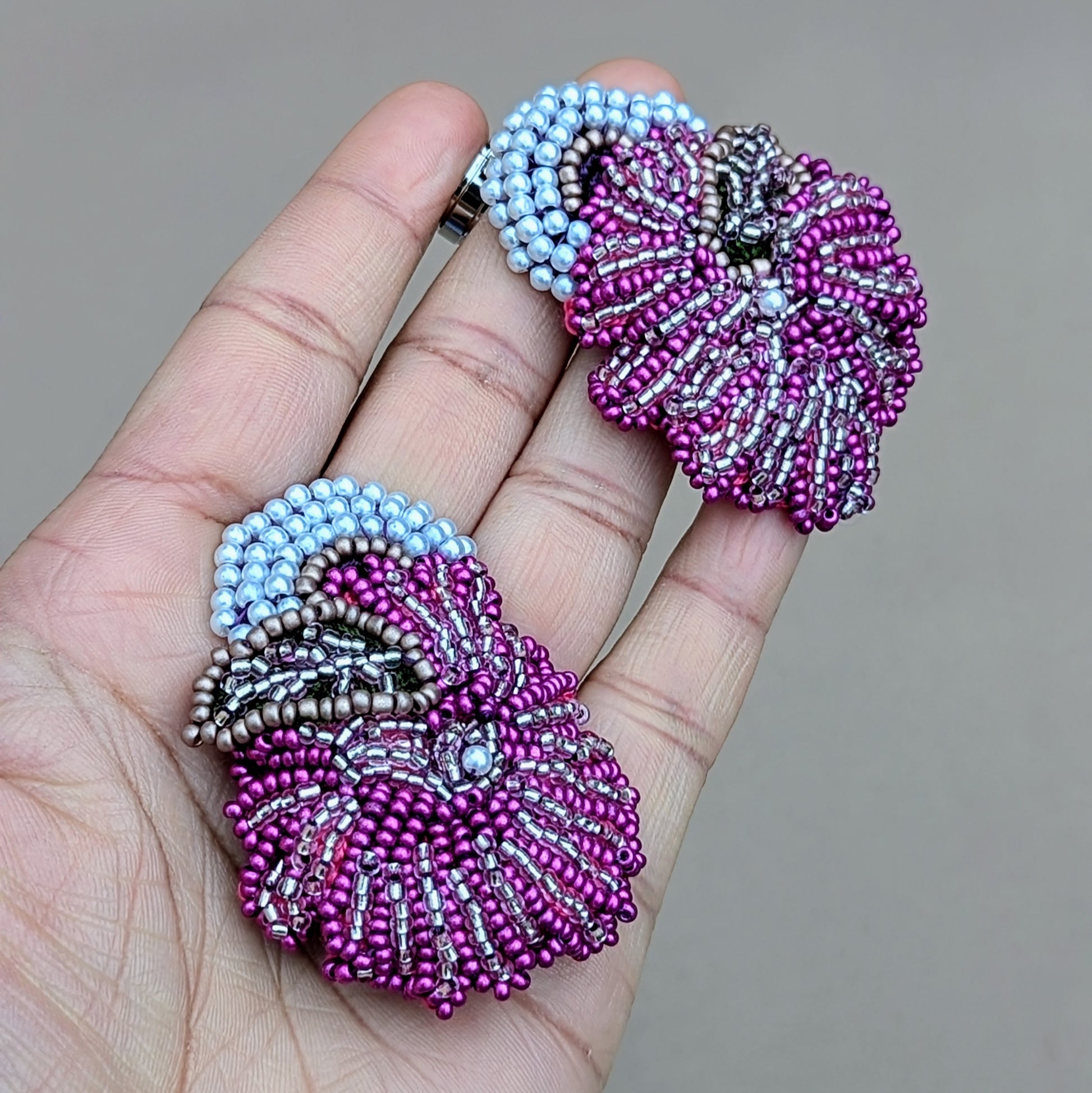 A hand holding a strikingly lightweight pair of intricately handcrafted Indian inspired earrings from Mayaani Jewellery, a sustainable UK artisan brand. The bold eye-catching design of these earrings features an exquisite magenta floral motif with silver and white pearl seed bead accents complimented perfectly with a stylish pair of heavy duty stainless steel earring clasps. 