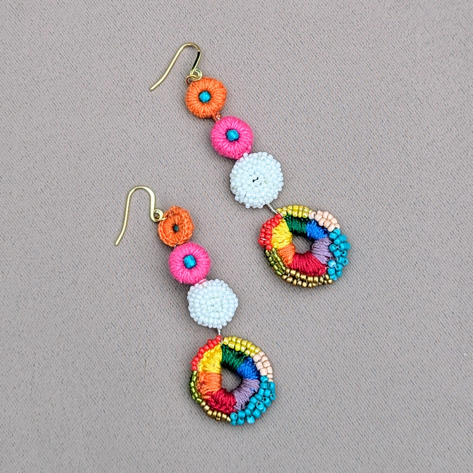 Adorn yourself with this vibrant pair of Indian-inspired rainbow earrings from Mayaani Jewellery UK. These earrings showcase a colourful rainbow crochet circle with intricate hand beadwork in red, green, yellow, peach, gold, and blue, highlighted by a white crochet and beaded frame. Pink and orange crochet accents complete the design, and the lightweight gold-plated stainless steel hooks make them perfect for joyful, all-day wear.