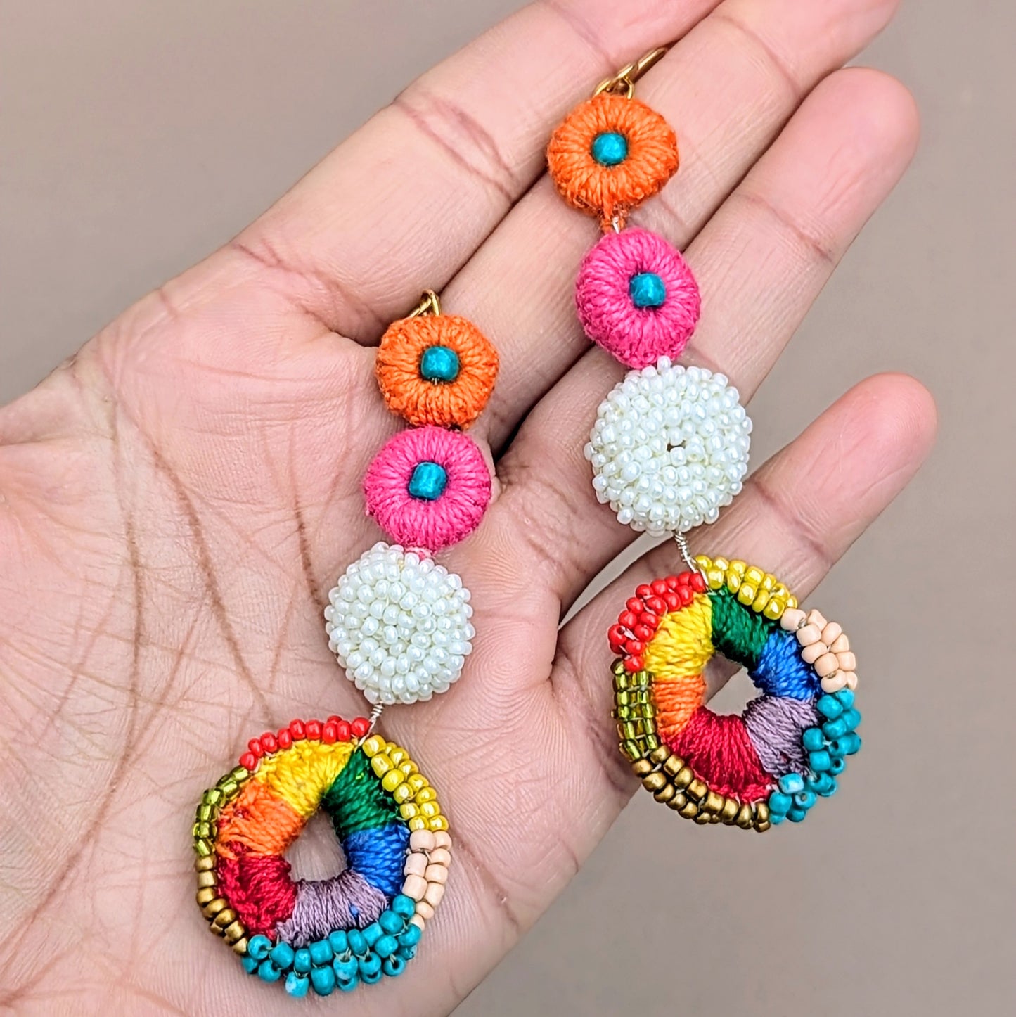 Introducing a pair of vibrant Indian-inspired rainbow earrings from Mayaani Jewellery UK. These earrings showcase a colourful rainbow crochet circle embellished with red, green, yellow, peach, gold, and blue hand beadwork, surrounded by a white crochet and beaded design. With pink and orange crochet accents, they are finished with lightweight gold-plated stainless steel hooks, ensuring effortless elegance.