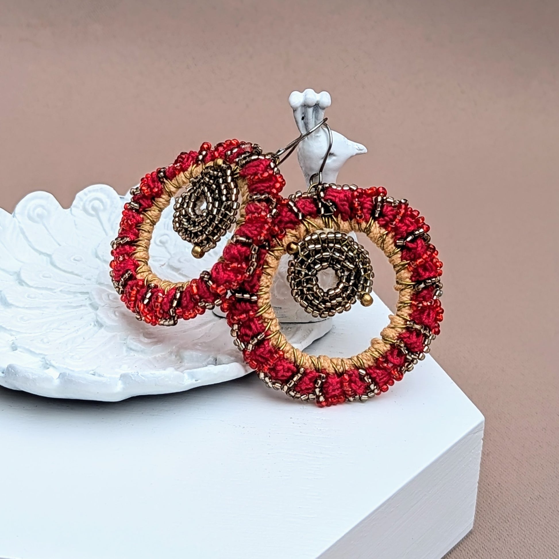 Featured on a white peacock trinket dish are an Indian inspired detailed pair of red hoop style earrings dripping in gold and silver accents. A unique pair exclusive to sustainable UK brand Mayaani Jewellery, each piece is precision handcrafted with the inclusion of high quality upcycled materials for a sumptuous blend of traditional artistry with a modern eco-friendly edge.