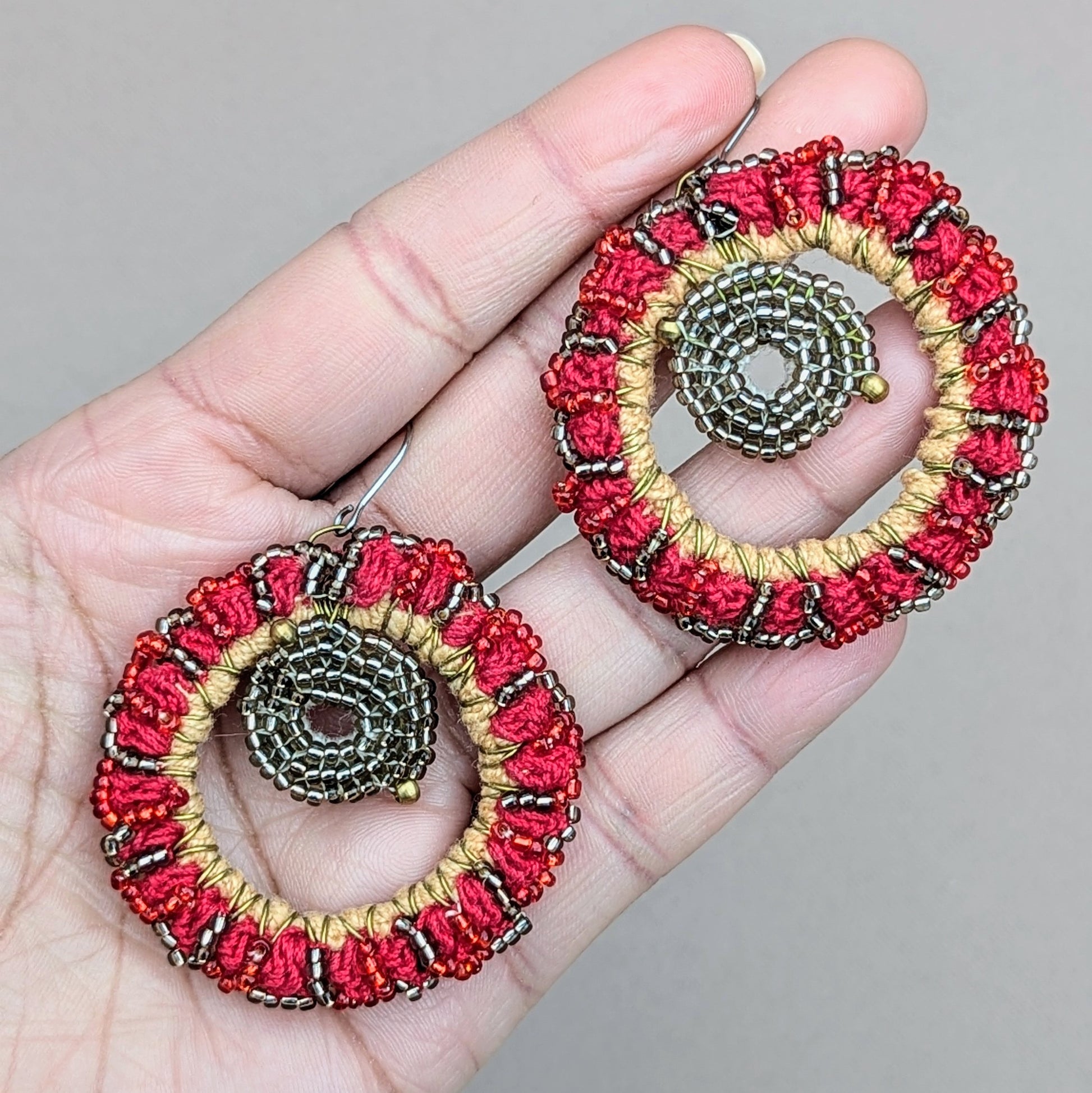 Hand holding a bold unique pair of artisan handcrafted Indian style earrings exclusively from the sustainable UK brand Mayaani Jewellery. The earrings feature a circular design with metallic silver and gold beaded centre framed by vibrant intricate red beadwork. With the inclusion of high quality upcycled materials this eco-conscious design creates a truly unique and sustainable accessory.