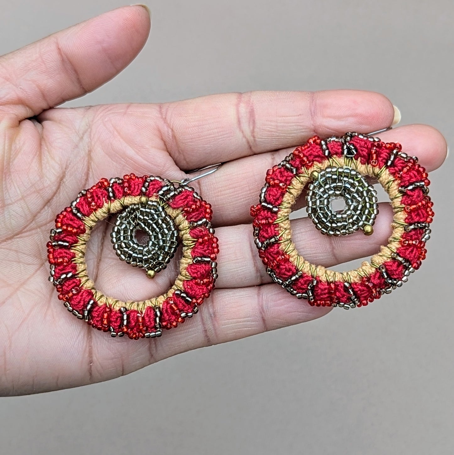 Hand holding a bold vibrant pair of red earrings featuring exquisite red and silver beadwork with gold accents in a circular design reminiscent of statement hoop style earrings. The high quality upcycled materials included in the design sing to the brand values of Mayaani Jewellery UK a sustainable handcrafted Indian wedding and occasion jewellery business.