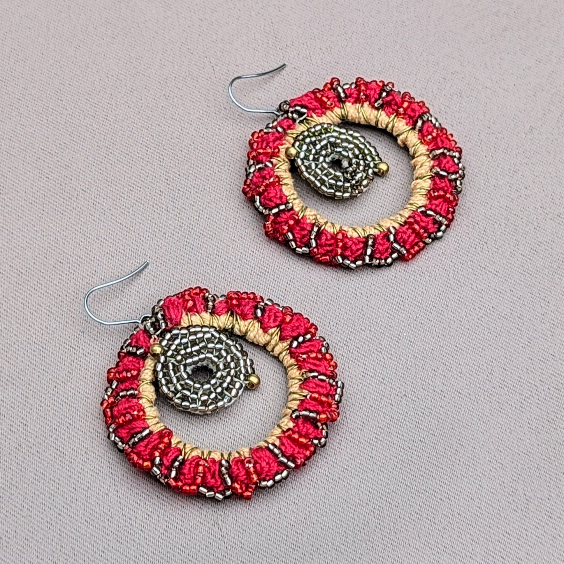 Experience the brilliance of style with sustainability in this standout pair of red earrings exclusive to sustainable UK brand Mayaani Jewellery. The earrings showcase high quality upcycled materials in a circular design with an intricate beaded centre in metallic silver, and gold tones reminiscent of a red hot sun. The outer ring is wrapped in golden thread, embellished with vibrant red accents and detailed beadwork, creating a bold and elegant contrast.