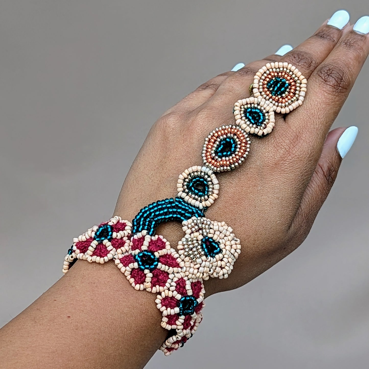 Presenting a unique designer Indian bridal hand bracelet, or haath phool, exclusive to Mayaani Jewellery, a sustainable UK brand. This stunning piece features intricate hand beadwork in shades of emerald green, peach, gold, and rose gold, arranged in a beautiful circular floral design. It gracefully connects the wrist to the finger with a delicate chain of beaded rings, showcasing detailed hand crochet and wire wrapping. Versatile and elegant, it can be worn as part of the matching bridal set or on its own.