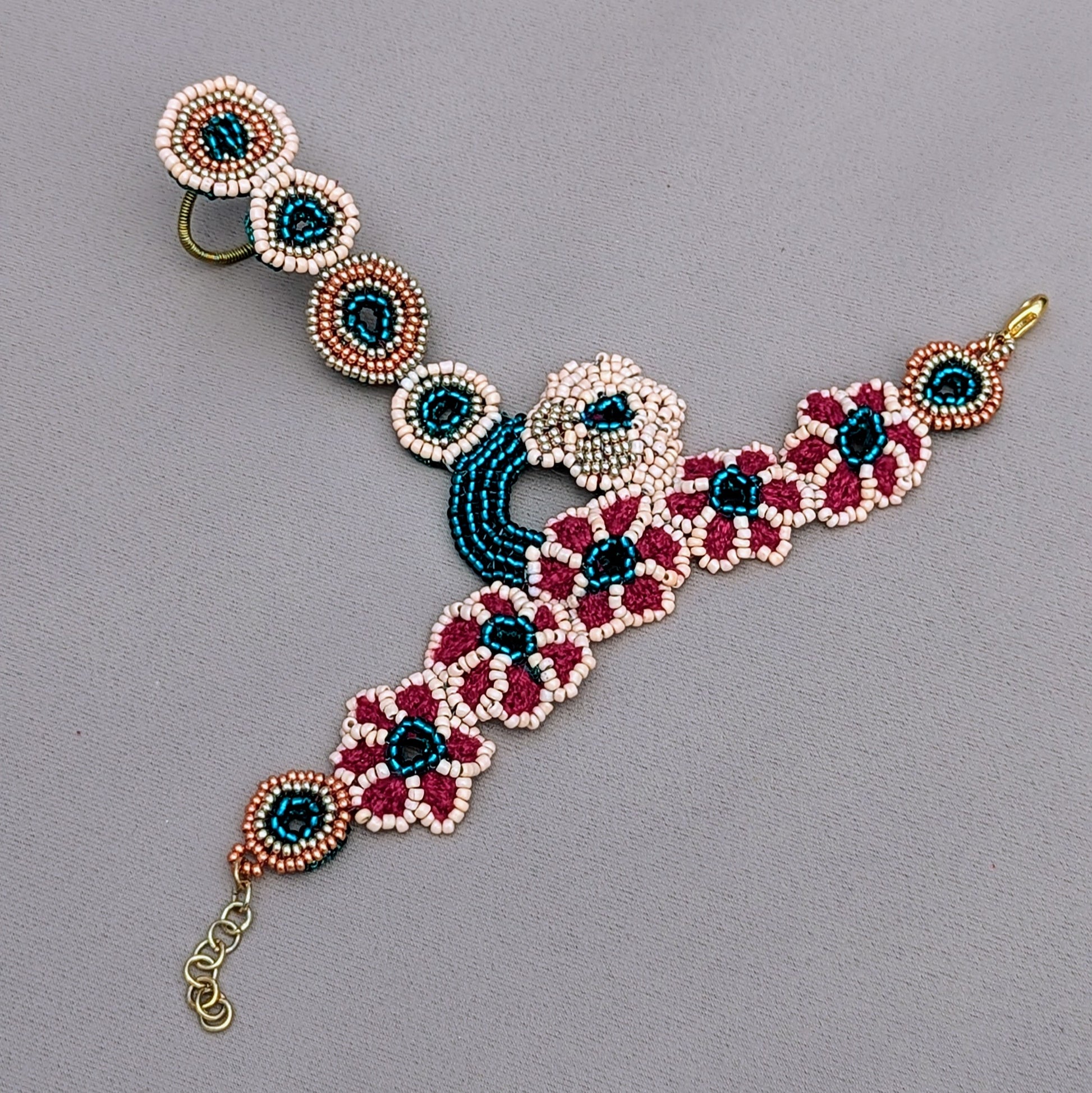 A distinctive designer Indian bridal hand bracelet, or haath phool, exclusively from Mayaani Jewellery, a sustainable UK brand. This elegant piece showcases intricate hand beadwork in vibrant emerald green, peach, gold, and rose gold tones, arranged in a charming circular floral design. It elegantly connects the wrist to the finger with a delicate chain of beaded rings, highlighting exquisite hand crochet and wire wrapping. Versatile and stylish, it can be paired with the matching bridal set or worn alone.