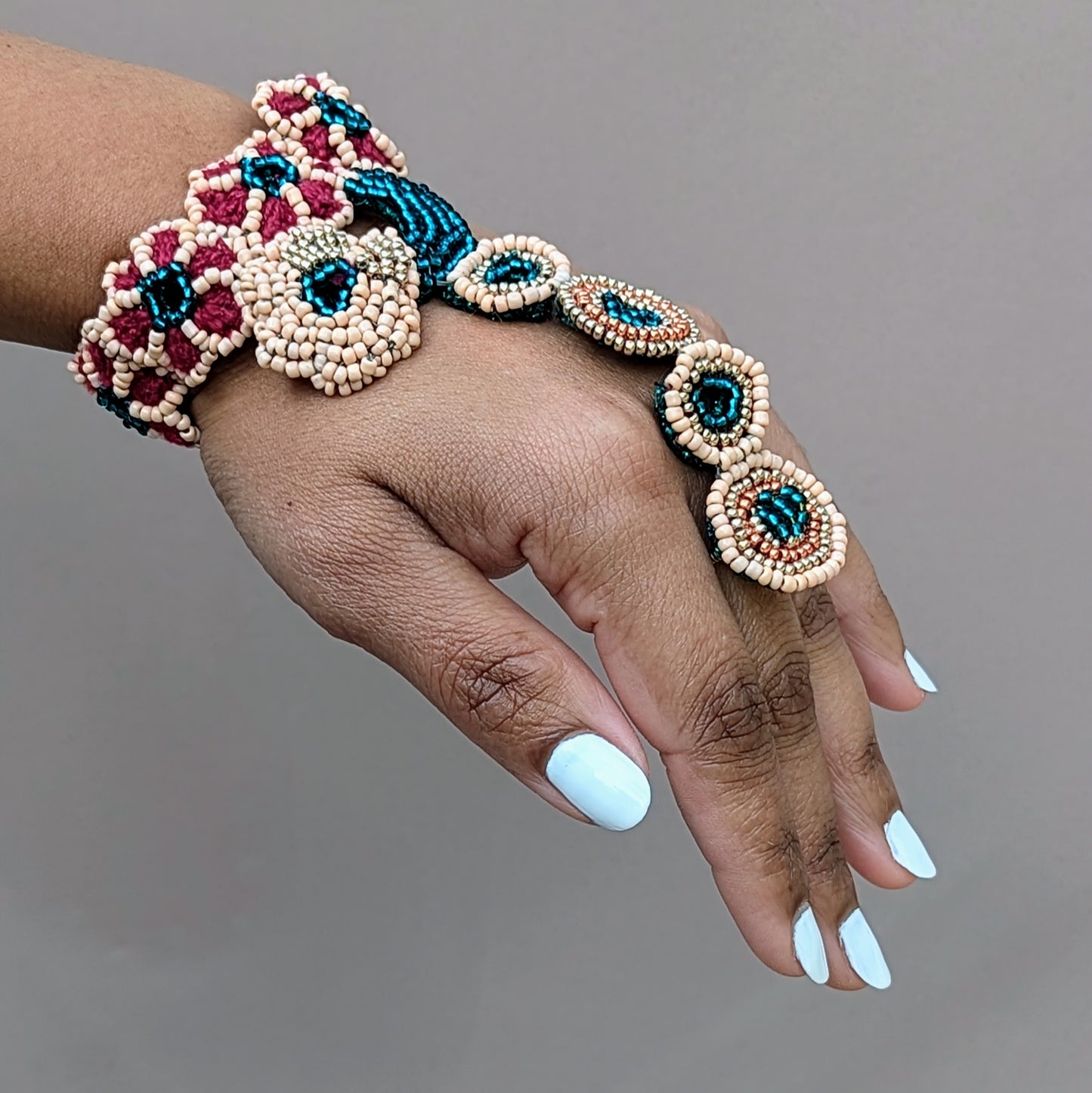 A unique designer Indian bridal hand bracelet, or haath phool, exclusive to Mayaani Jewellery, a sustainable UK brand. This piece features intricate hand beadwork in emerald green, peach, gold, and rose gold tones, beautifully arranged in a circular floral design. It elegantly connects the wrist to the finger with a delicate chain of beaded rings, highlighting detailed hand crochet and wire wrapping. Versatile in nature, it can be paired with the matching bridal set or worn alone for special occasions.