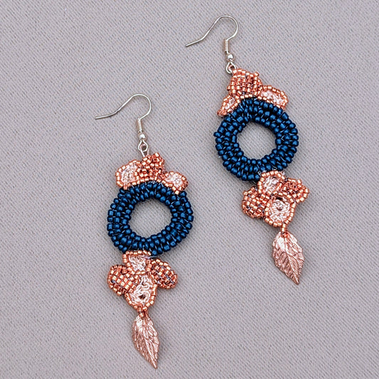 A striking pair of handcrafted earrings featuring a large circular blue beaded centerpiece surrounded by floral motifs made of copper Japanese Miyuki seed beads. Gold leaf accents and delicate hand-crochet and beadwork enhance the design, while rose gold-plated hooks add a luxurious touch. Created by sustainable UK brand Mayaani Jewellery, these Indian-inspired earrings blend deep blue glass seed beads with premium materials, embodying a perfect balance of artistry and sustainable fashion. 