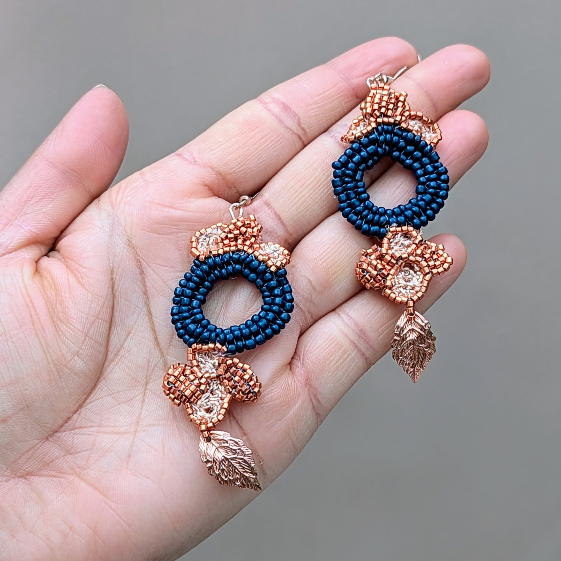 A pair of unique, handcrafted earrings showcasing a large circular blue beaded centrepiece, adorned with floral motifs made from copper Japanese Miyuki seed beads. Gold leaf accents and intricate hand-crochet and beadwork enhance the design, finished with elegant rose gold-plated hooks. These Indian-inspired earrings by Mayaani Jewellery blend deep blue glass seed beads and premium materials, perfectly merging artistry with sustainable fashion.