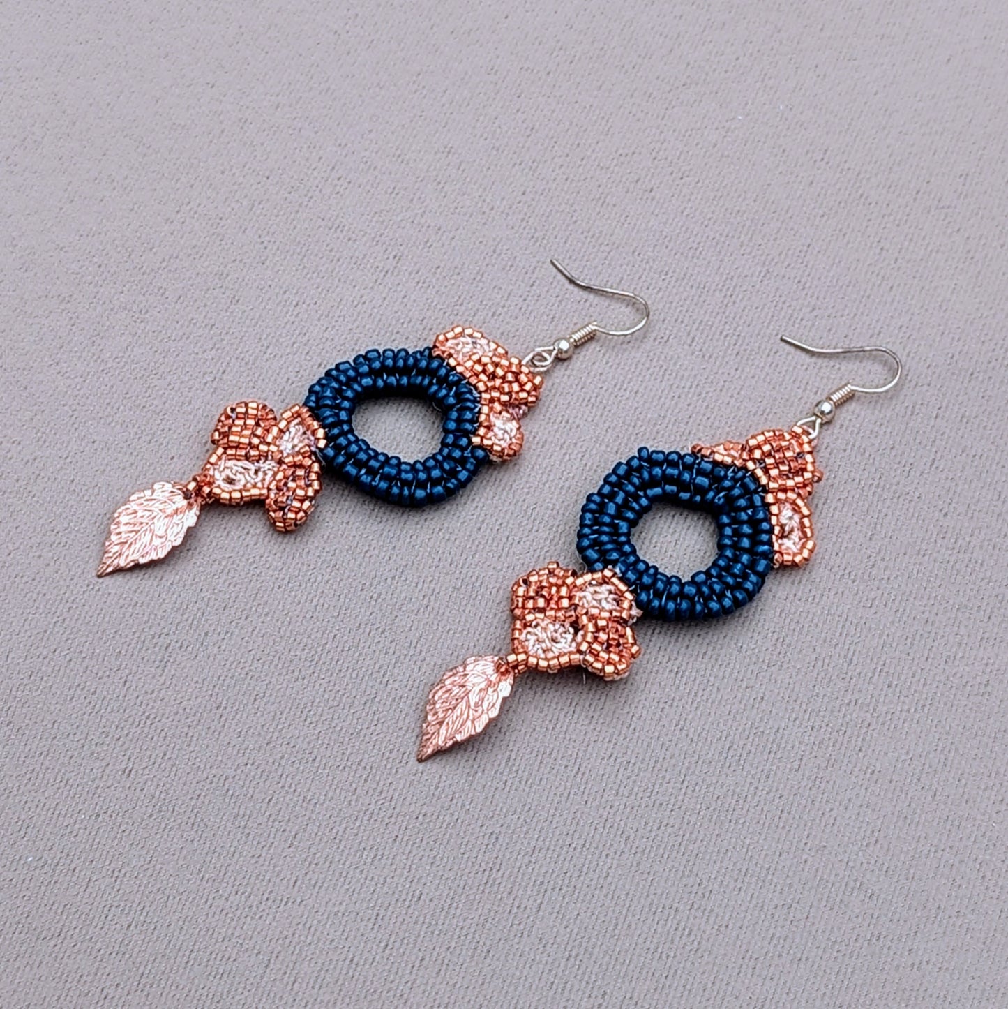  A striking pair of earrings showcasing a large circular blue beaded centrepiece, surrounded by floral motifs crafted from copper Japanese Miyuki seed beads. Enhanced with gold leaf accents, the earrings feature delicate hand-crochet and beadwork, complemented by luxurious rose gold-plated hooks. Handcrafted by sustainable UK brand Mayaani Jewellery, these earrings merge traditional Indian design with modern artistry. Dimensions: max width 2.7cm, approx. length 7cm.