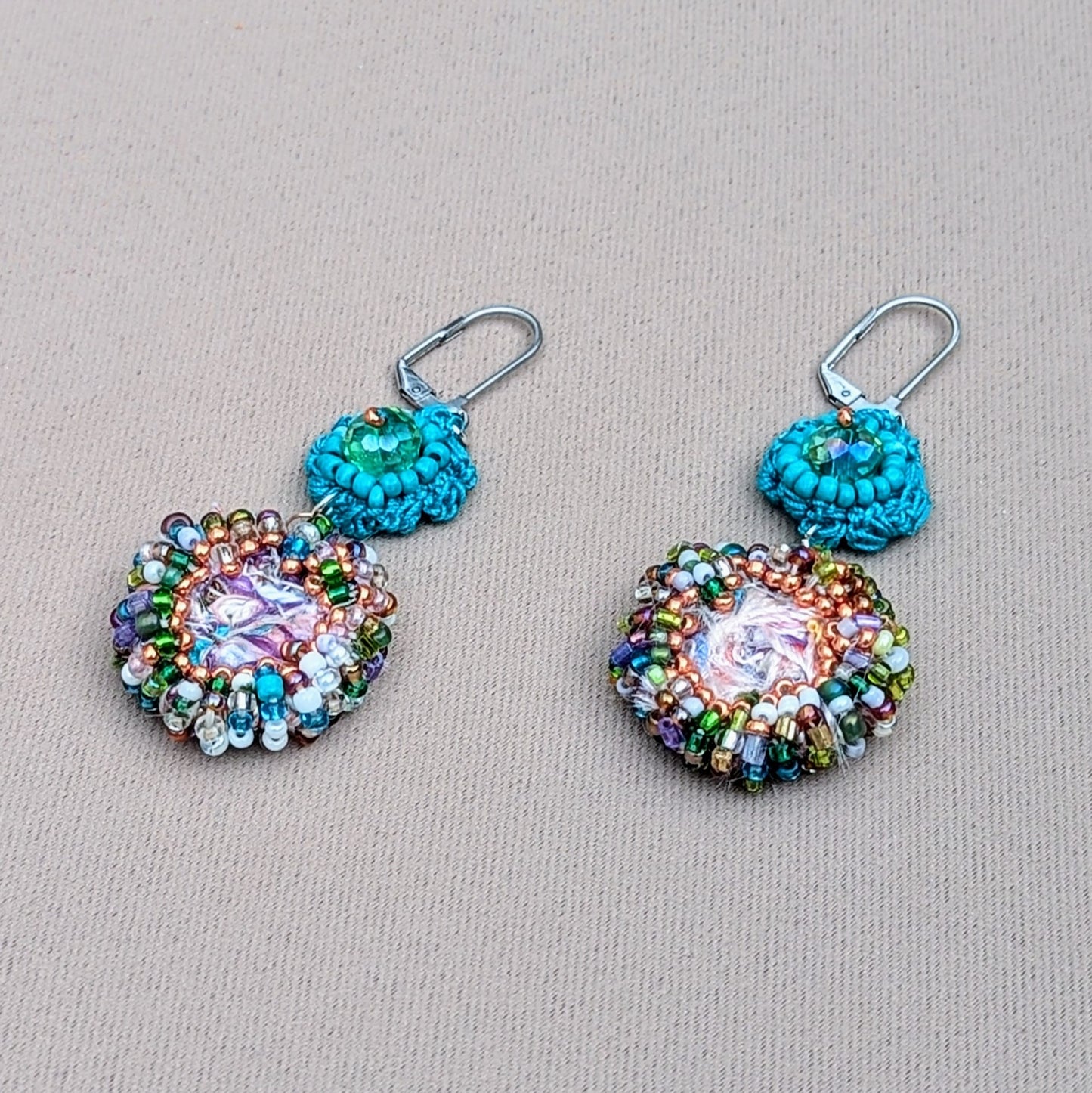 A stunning pair of sustainable scrap bead earrings by Mayaani Jewellery UK, reflecting the beauty of crystal-clear ocean waters and Indian design. Each earring features a large handcrafted circular scrap bead made from jewellery-making remnants, adorned with high-quality upcycled seed beads in purple, rose gold, green, white, and blue. A hand-crocheted turquoise flower motif with an upcycled Swarovski-style bicone bead adds a finishing touch. Perfect for weddings and special occasions, as well as everyday.